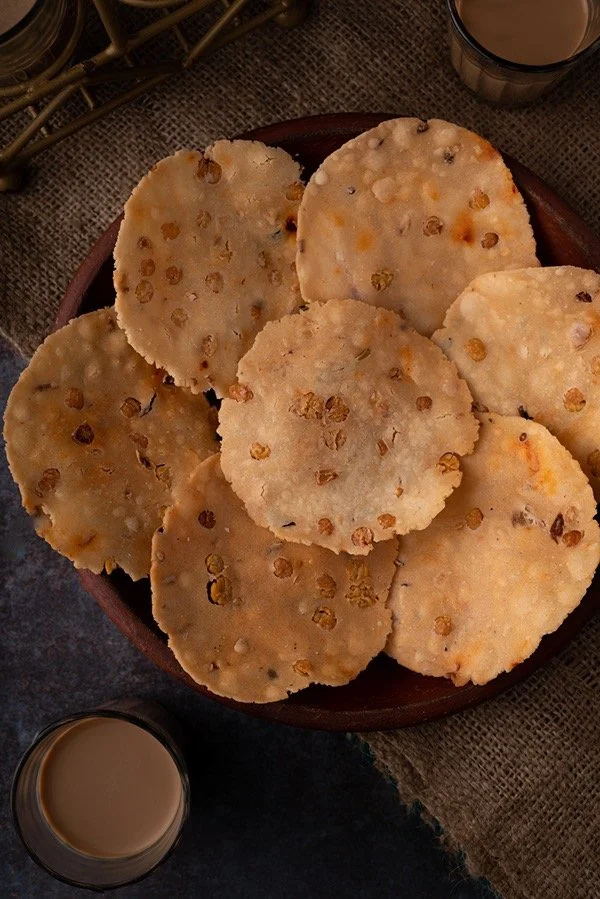 A wooden plate containing several round, crispy flatbread chapatis with specks of seasoning, placed on a textured cloth surface with glasses of chai tea nearby.