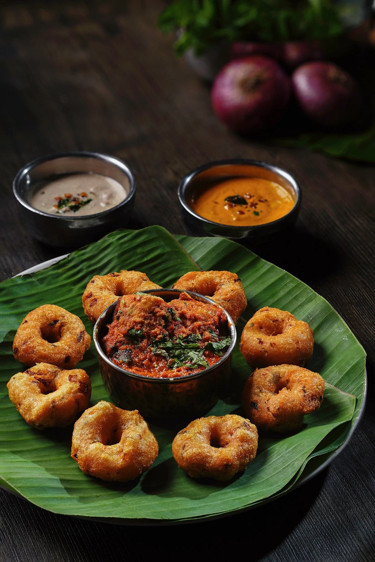 A plate of Medu Vada, a South Indian savoury snack, arranged on banana leaf, served with spicy chutney in a small bowl, accompanied by two dipping sauces and red onions in the background.