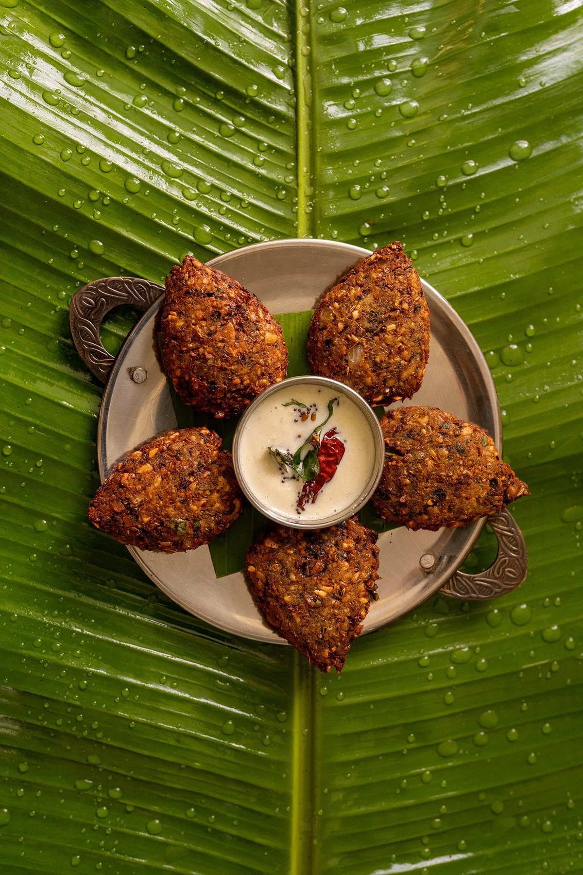 A plate of five stuffed Indian eggplants, called brinjal or aubergine, with a small bowl of yogurt-based sauce garnished with chili and curry leaves, placed on a large, wet, green banana leaf.