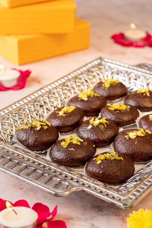 A silver tray filled with chocolate ladoos garnished with chopped pistachios, situated on a floral-themed table with small candles and decorative yellow boxes in the background.