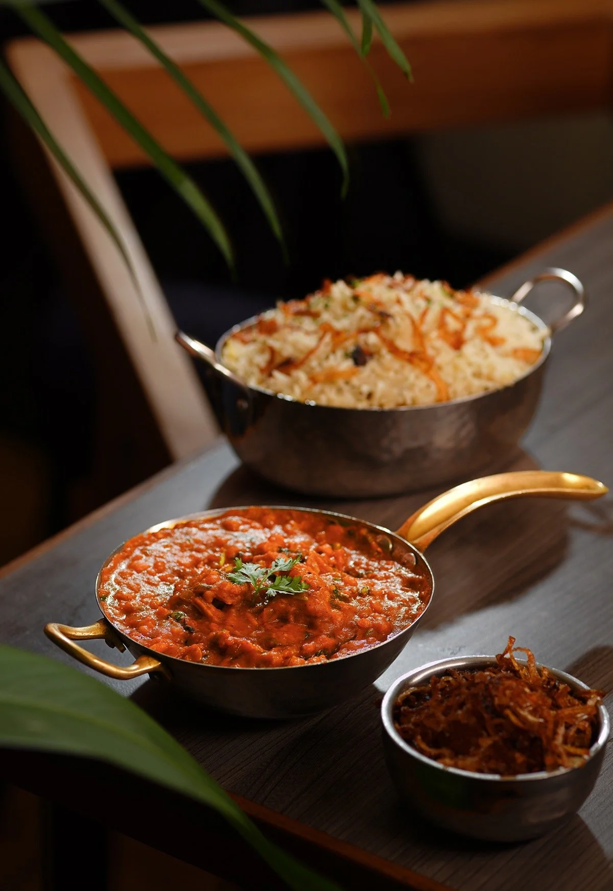 Three bowls of Indian dishes placed on a wooden surface, with a green plant in the foreground. The dishes include rice with vegetables, a spicy curry garnished with herbs, and a side dish of shredded vegetables or pickles.