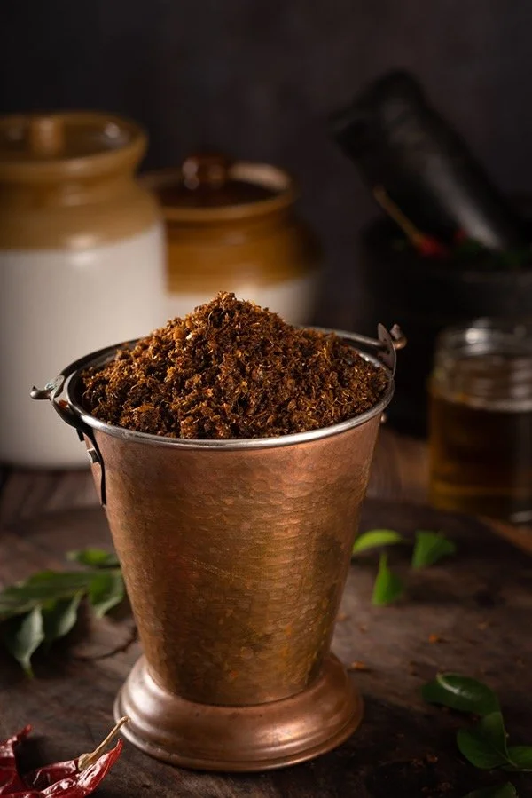 A copper vessel filled with ground spices, possibly used for cooking or seasoning, placed on a wooden surface surrounded by leaves and kitchen jars in the background.