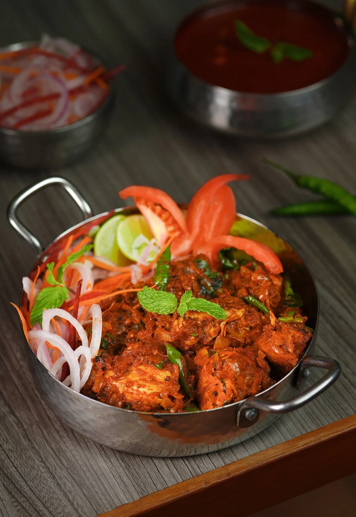 Dish of cooked chicken in spicy red sauce garnished with mint, served with sliced tomatoes, lime wedges, and fresh green chili peppers, on a wooden surface.