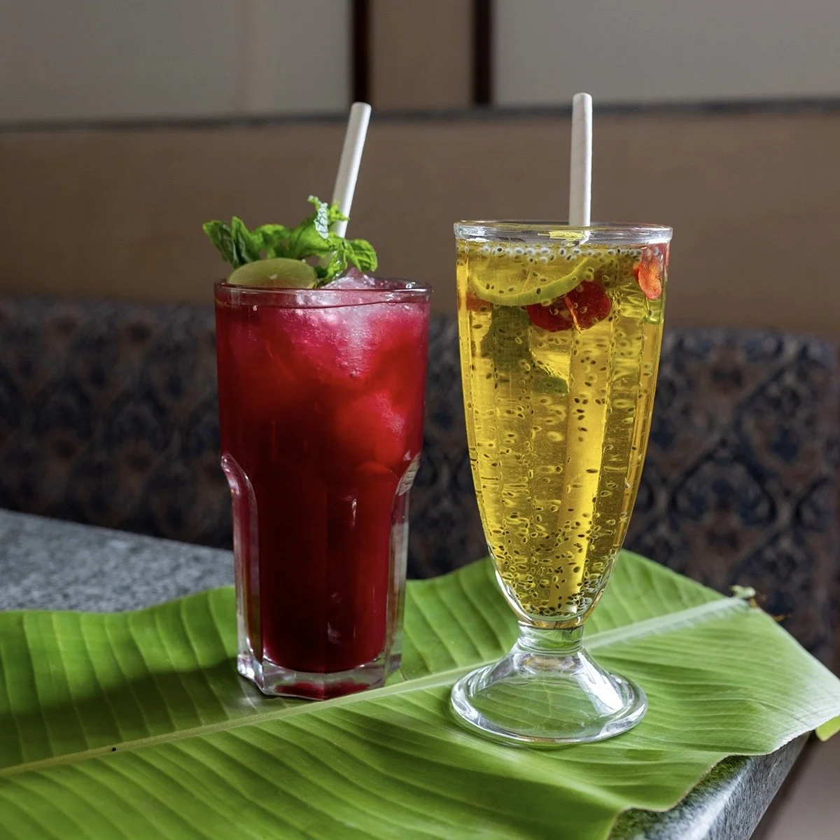 Two tropical drinks, one red with ice and garnished with grapes and mint, and one yellow with bubbles and fruit slices, placed on a green banana leaf on a table.