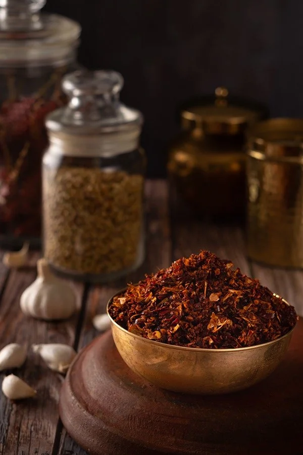A metal bowl filled with crushed dried red chili peppers on a wooden surface, with jars of spices and garlic cloves in the background.