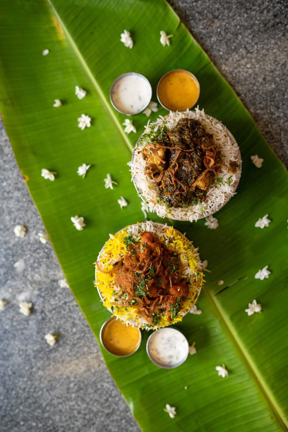 Two bowls of Indian rice dishes placed on a green banana leaf with small bowls of sauces and sprinkled flower petals.
