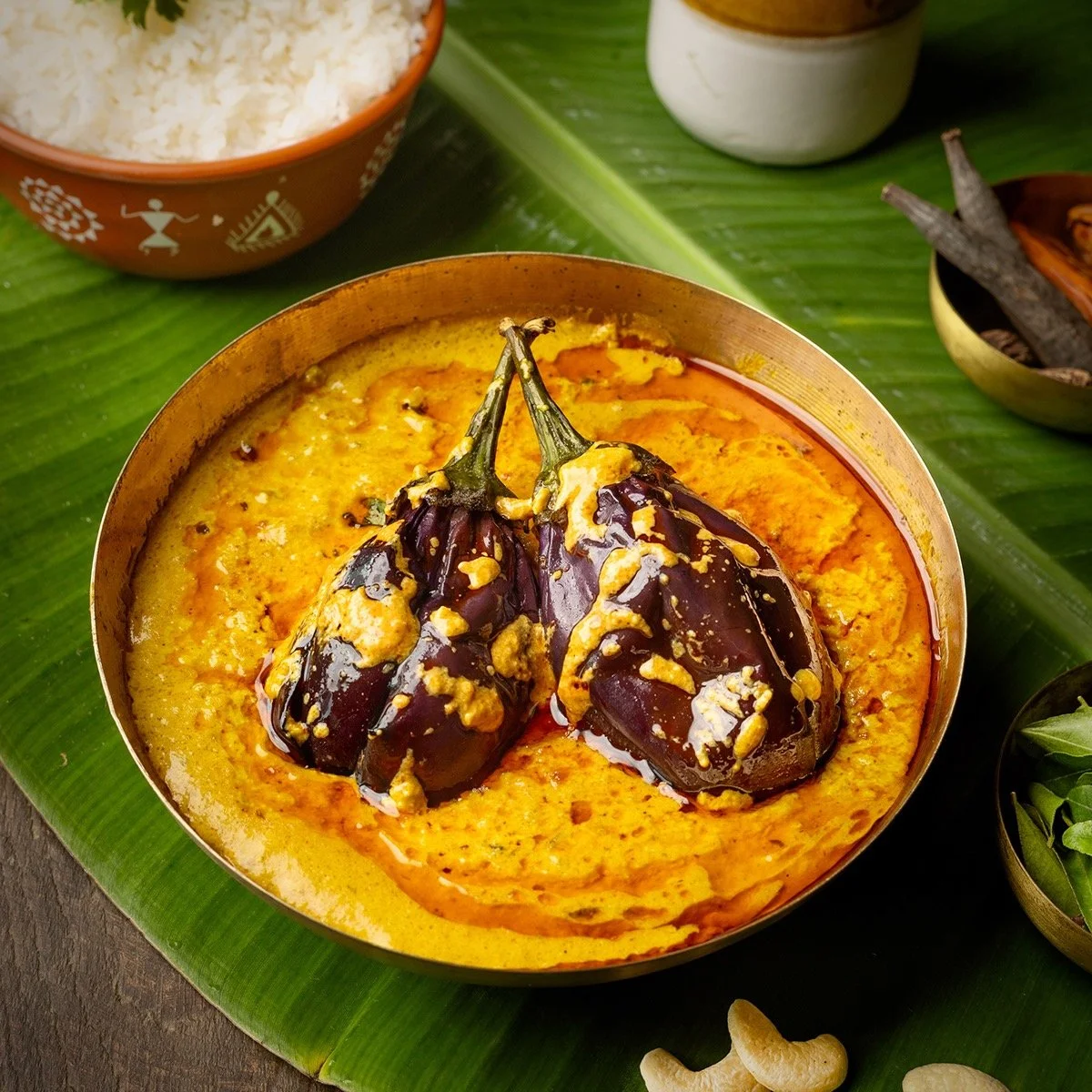 A traditional Indian dish with two dark purple eggplants in a yellow curry, garnished with a yellow sauce, served in a decorative bowl on a green banana leaf, with other bowls of rice and spices in the background.