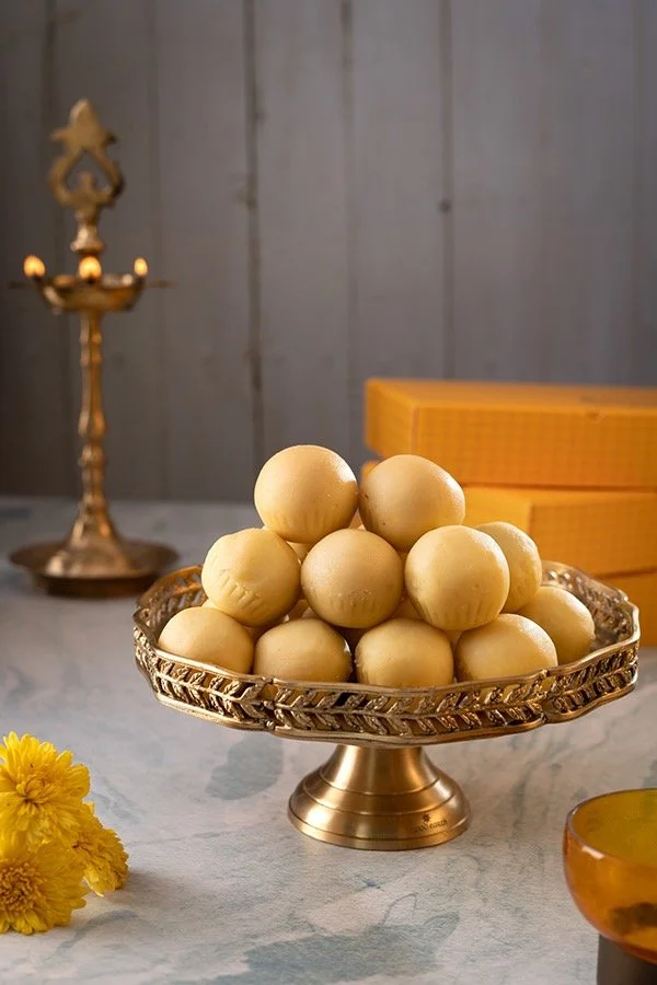 A silver decorative platter filled with round, pale yellow sweets, likely Indian confections, on a white table with yellow boxes, a yellow flower, and an ornate gold candle holder in the background.