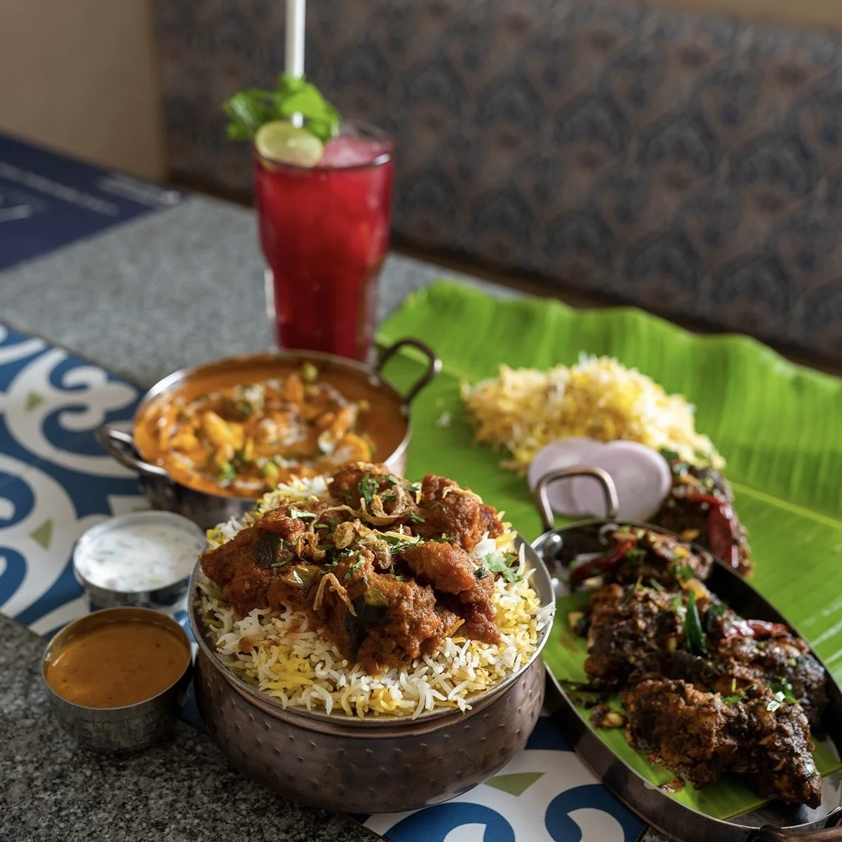 A traditional Indian meal with curry, rice, and grilled meats served on a banana leaf, accompanied by a glass of red drink with lime and mint.