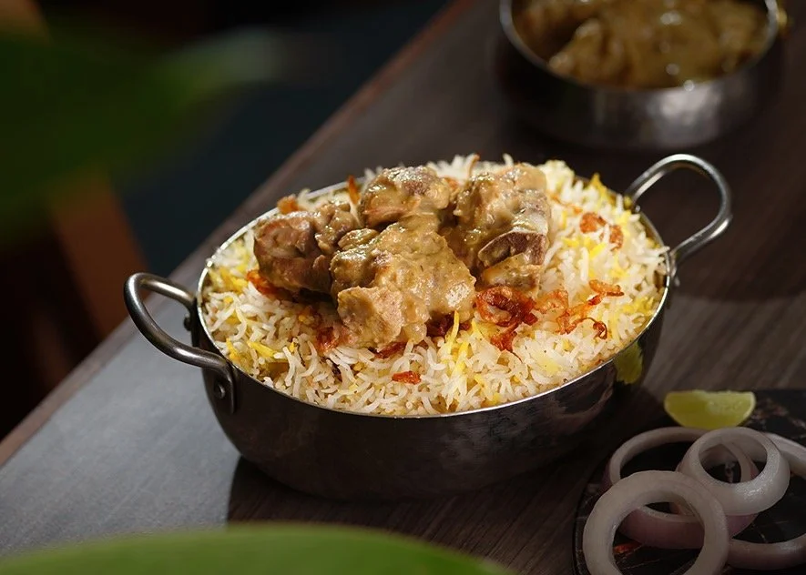 A bowl of white rice topped with some cooked meat in gravy, garnished with fried onions, and served in a metallic bowl with handles. There is a lemon wedge, sliced onions, and another dish in the background.