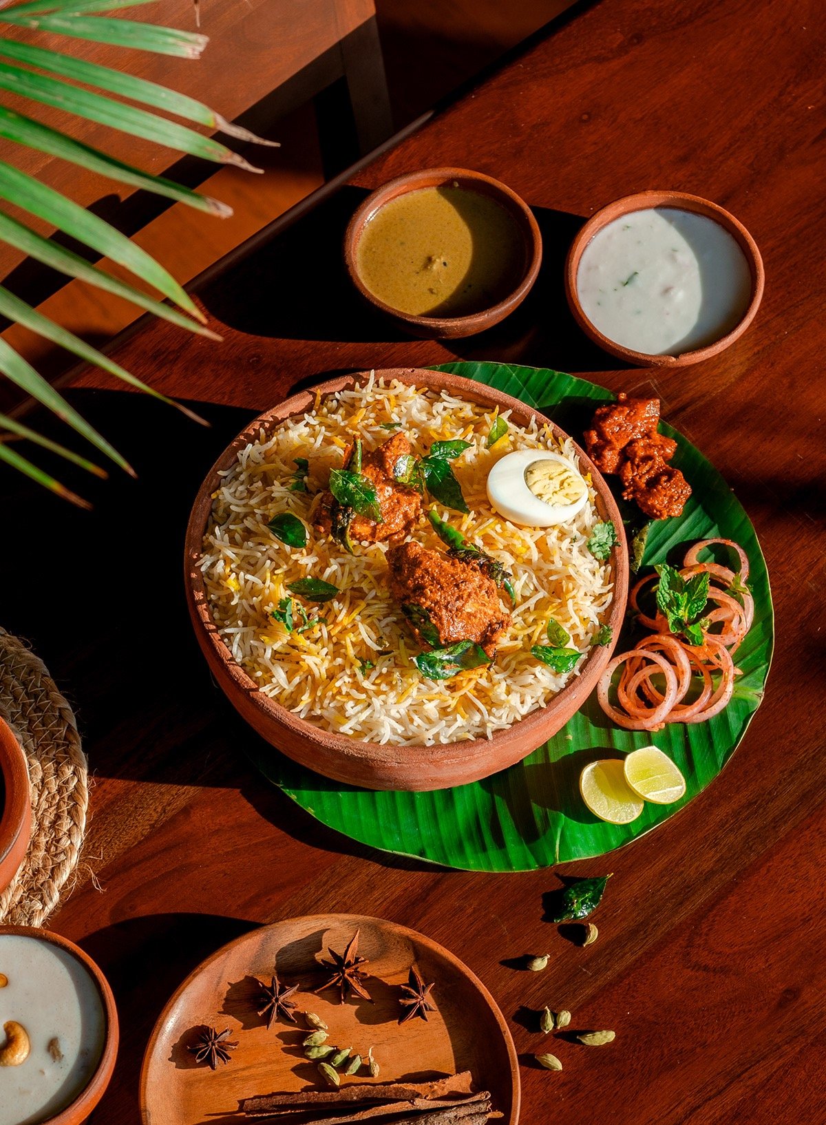 A traditional Indian meal featuring a large bowl of spices and herbs rice, half a boiled egg, and garnishes like lemon slices, onions, and herbs, with sides of mint yogurt and green chutney, on a wooden table with spices.