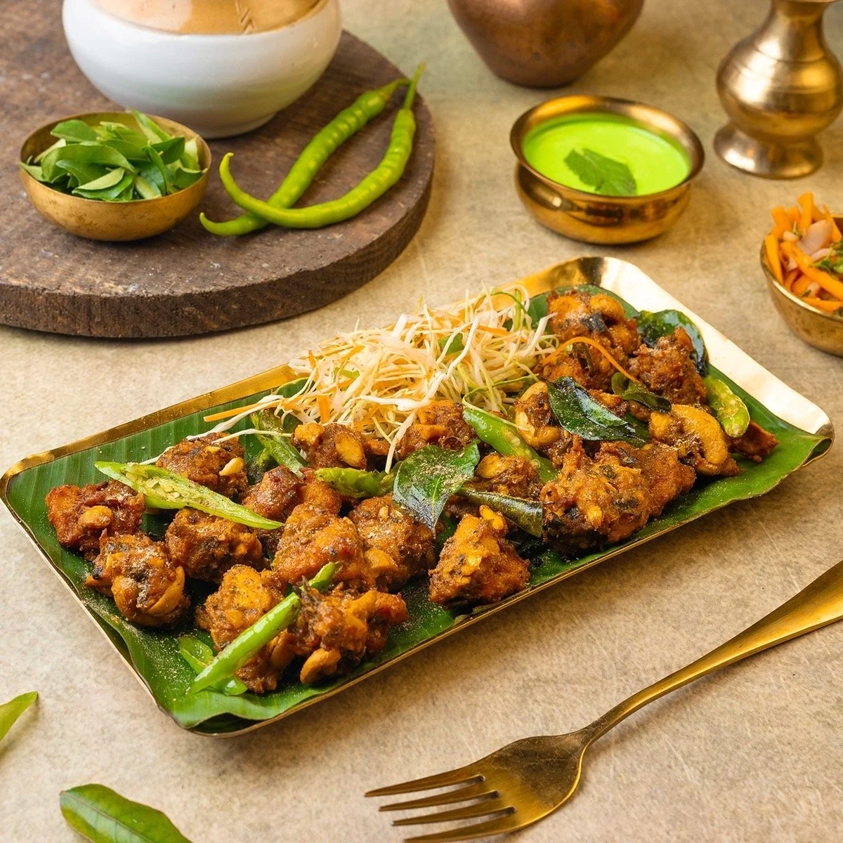 A plate of Indian-style chicken curry garnished with green chili and served with shredded vegetables. Surrounding dishes include bowls of green chutney, salad, and spices.