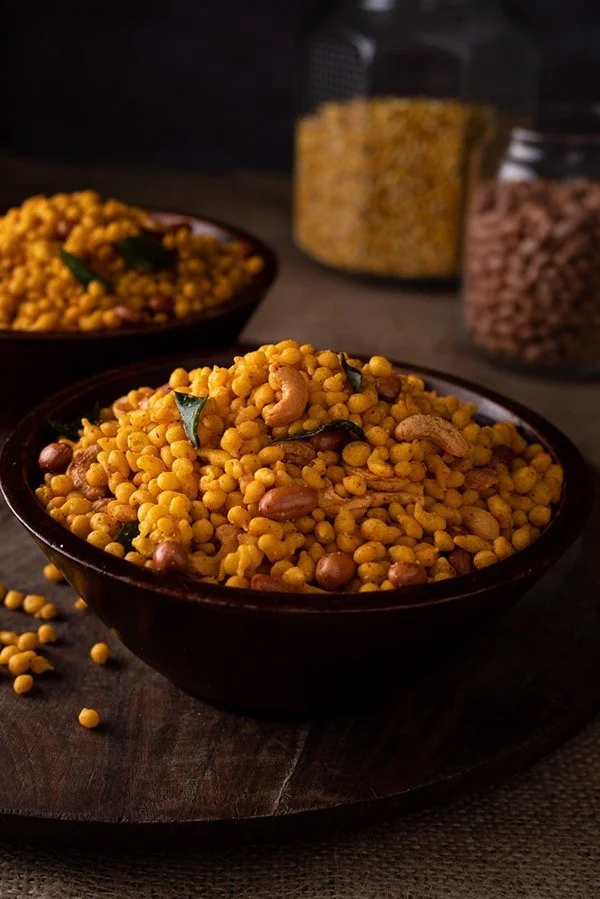 A bowl of Indian snack mixture with yellow puffed rice, nuts, and spices on a dark wooden surface, with jars of more snack ingredients in the background.