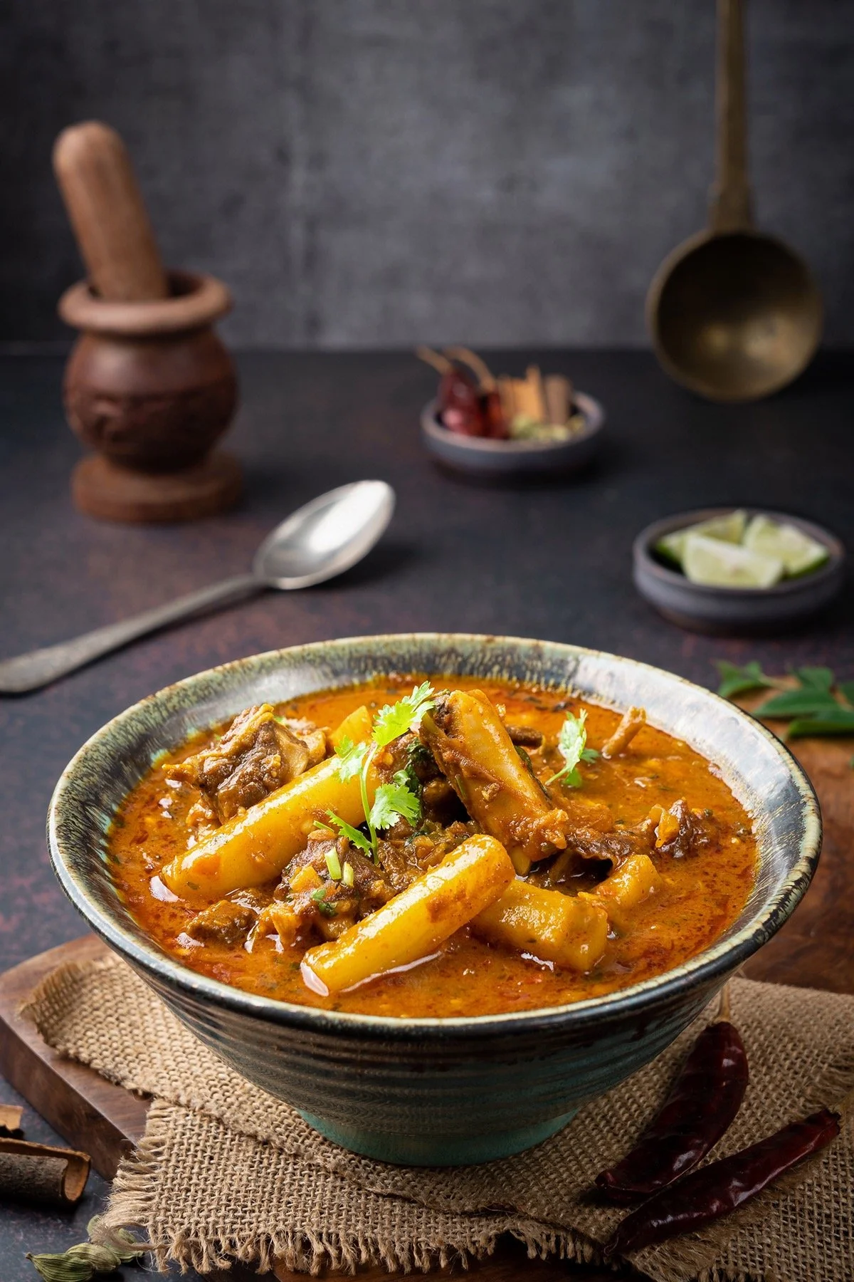 A bowl of Indian curry with potatoes, garnished with cilantro, on a textured cloth. In the background, there are small bowls with lime wedges and spices, and a few dried red chilies on the table.