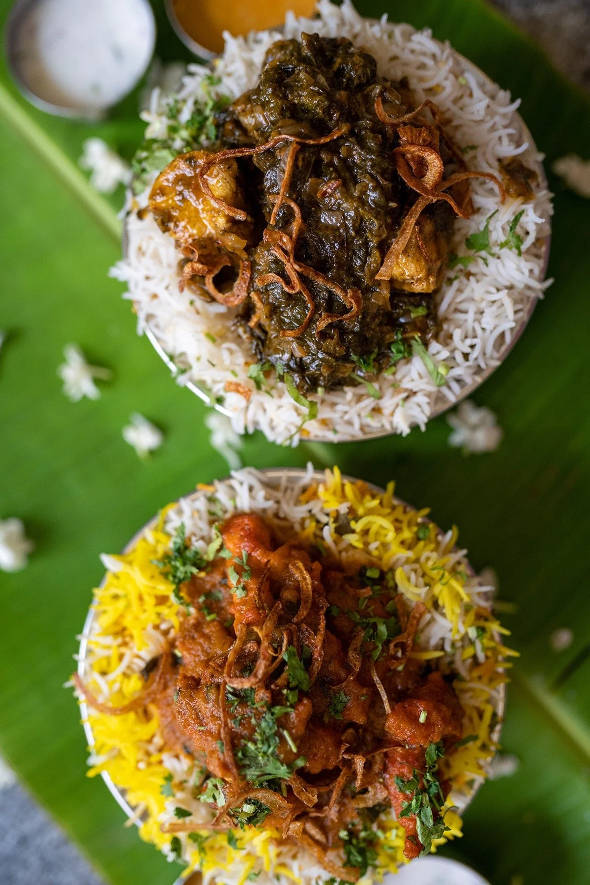 Two bowls of Indian dishes on a banana leaf, topped with fried onions, garnished with cilantro, with white rice and yellow rice underneath, and small cups of sauce or yogurt on the side.