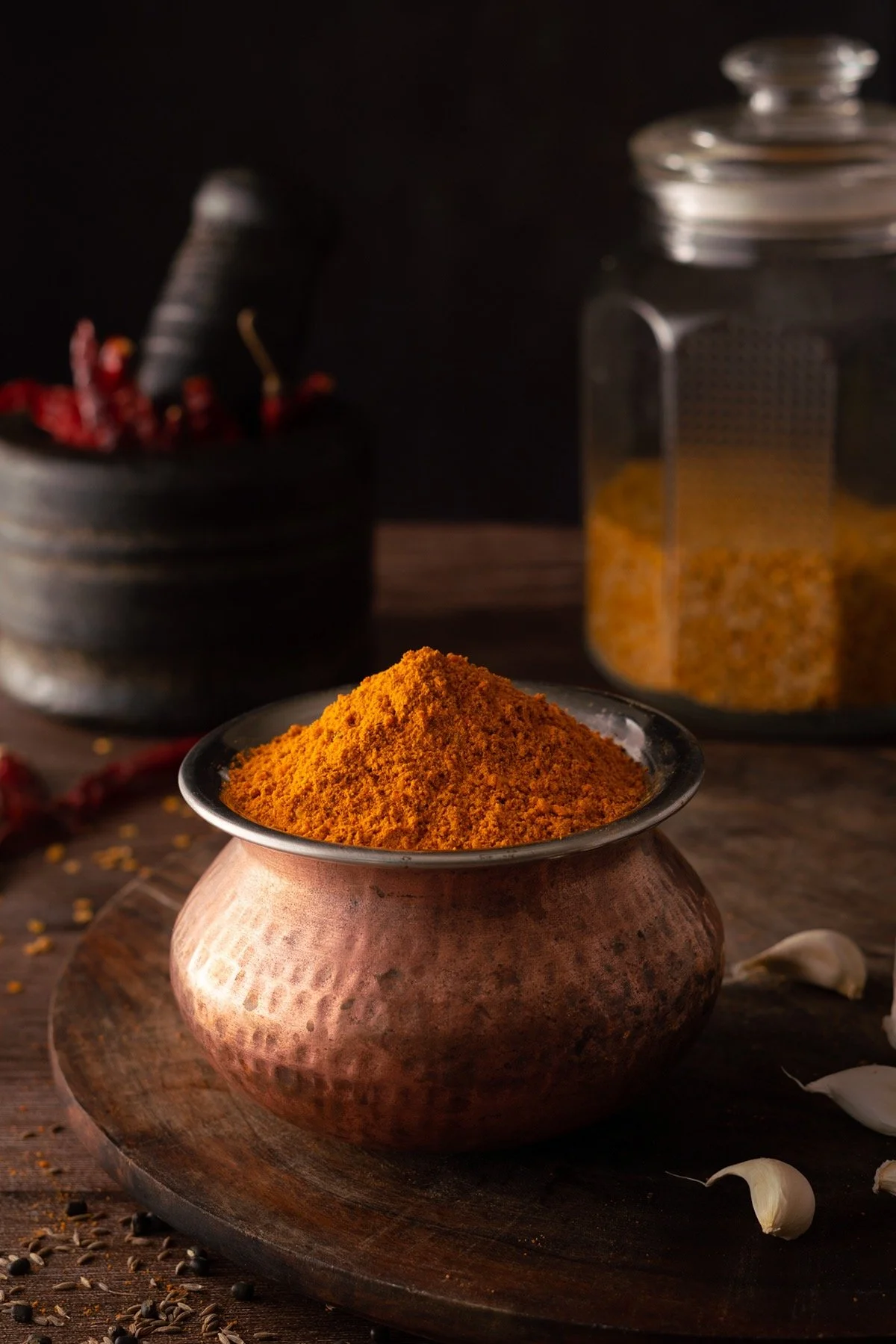 A bowl of bright orange turmeric powder on a wooden surface, with garlic cloves, black peppercorns, and containers of spices in the background.