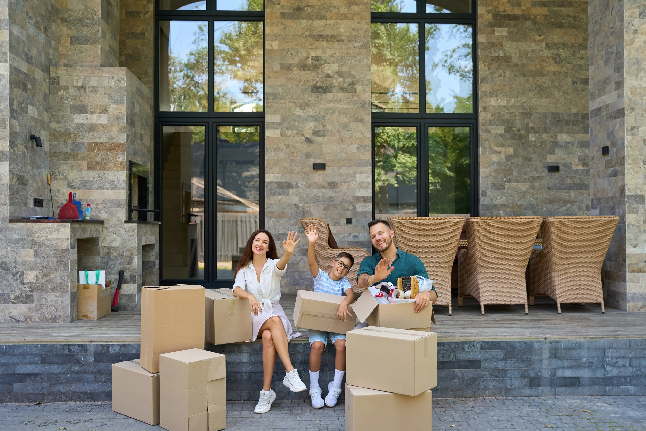 Happy family sitting on a porch surrounded by moving boxes, waving at the camera after moving into a new home.