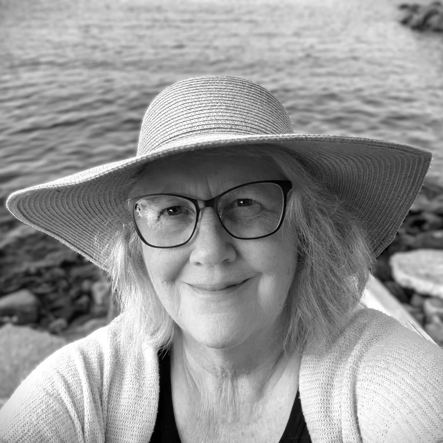A smiling woman wearing glasses and a large sunhat takes a selfie near a body of water with rocks in the background.