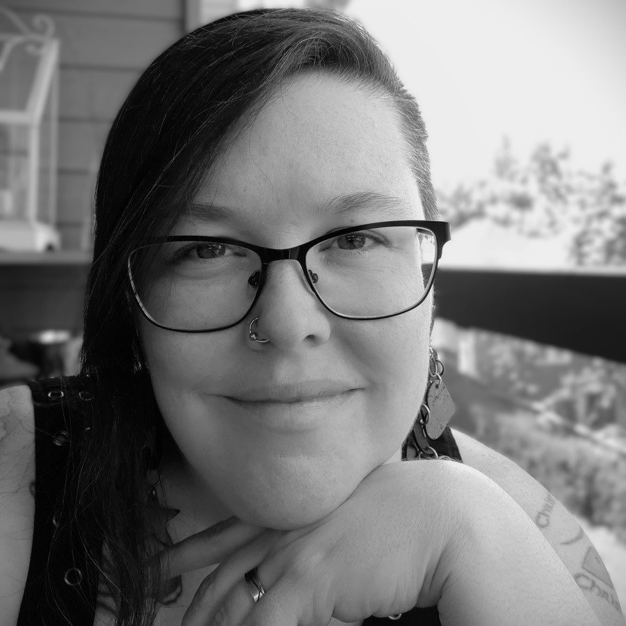 Black-and-white photo of a woman with glasses, nose piercings, and earrings, smiling and resting her chin on her hand, with a blurred outdoor background.