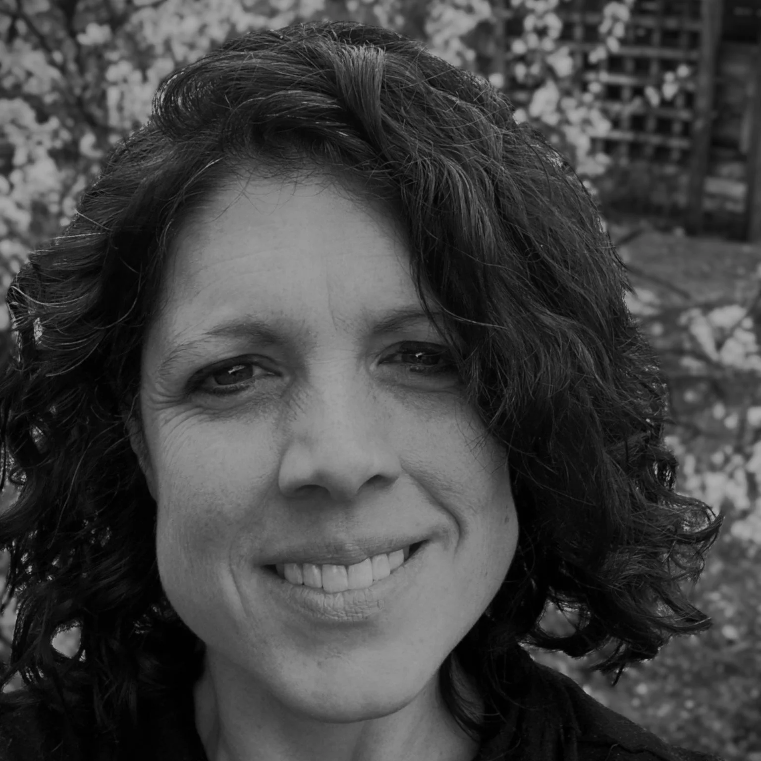 Close-up of a woman with curly hair, smiling, outdoors in front of flowering trees.