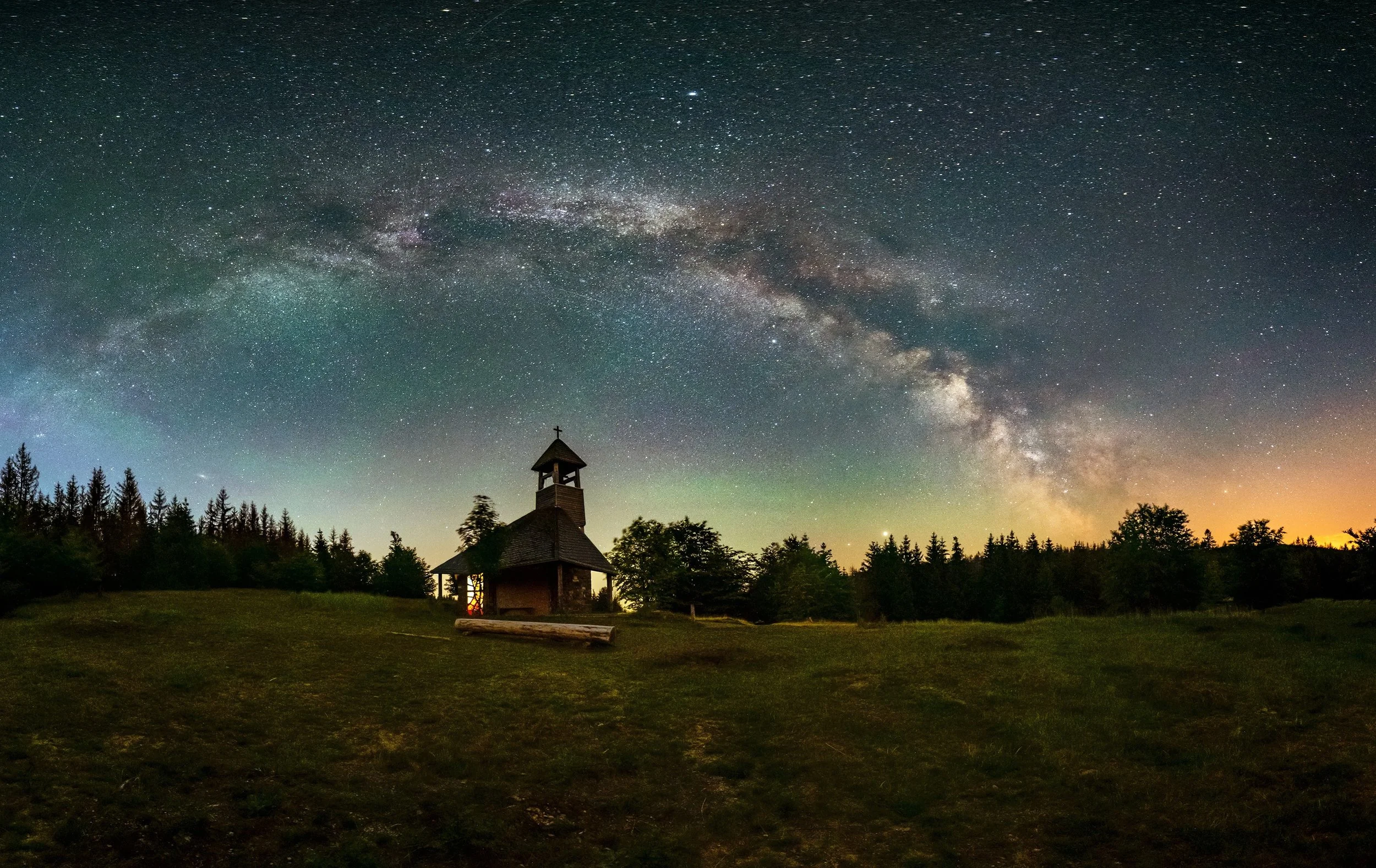 Night sky with the Milky Way galaxy arching over a silhouette of a small wooden church and trees.