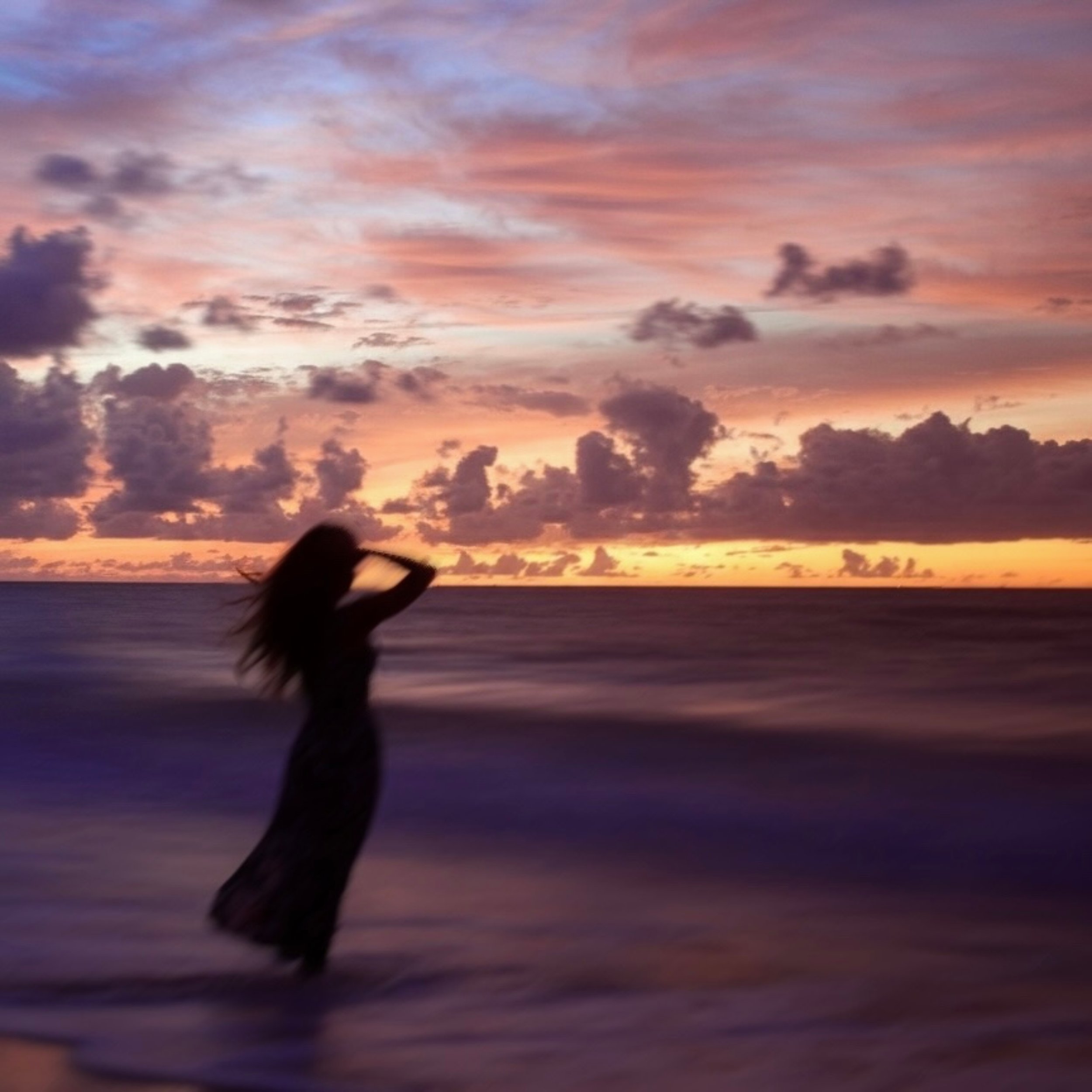 A silhouette of a woman standing in the ocean at sunset, with colorful pink, purple, and orange clouds in the sky.