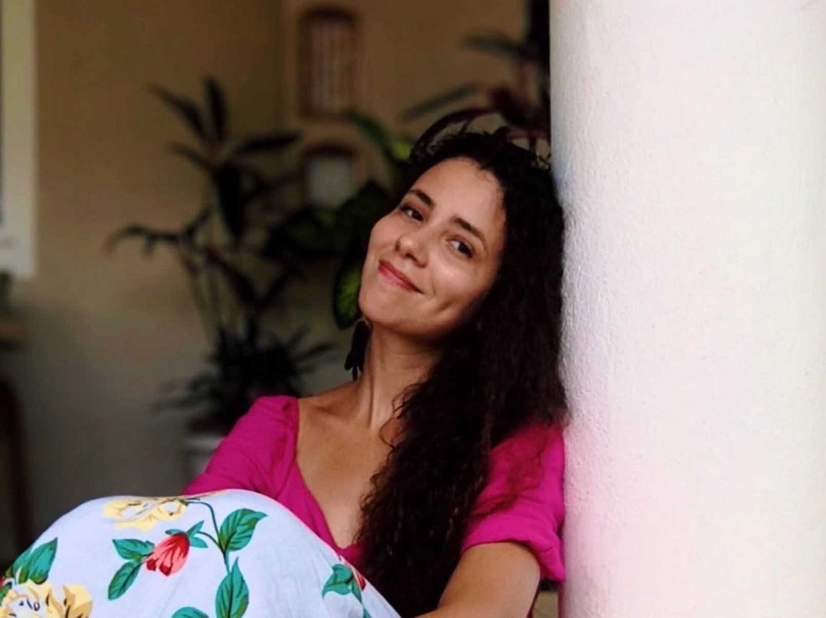 A woman with dark, curly hair and a pink shirt sitting against a white wall, smiling at the camera with plants and a picture frame in the background.