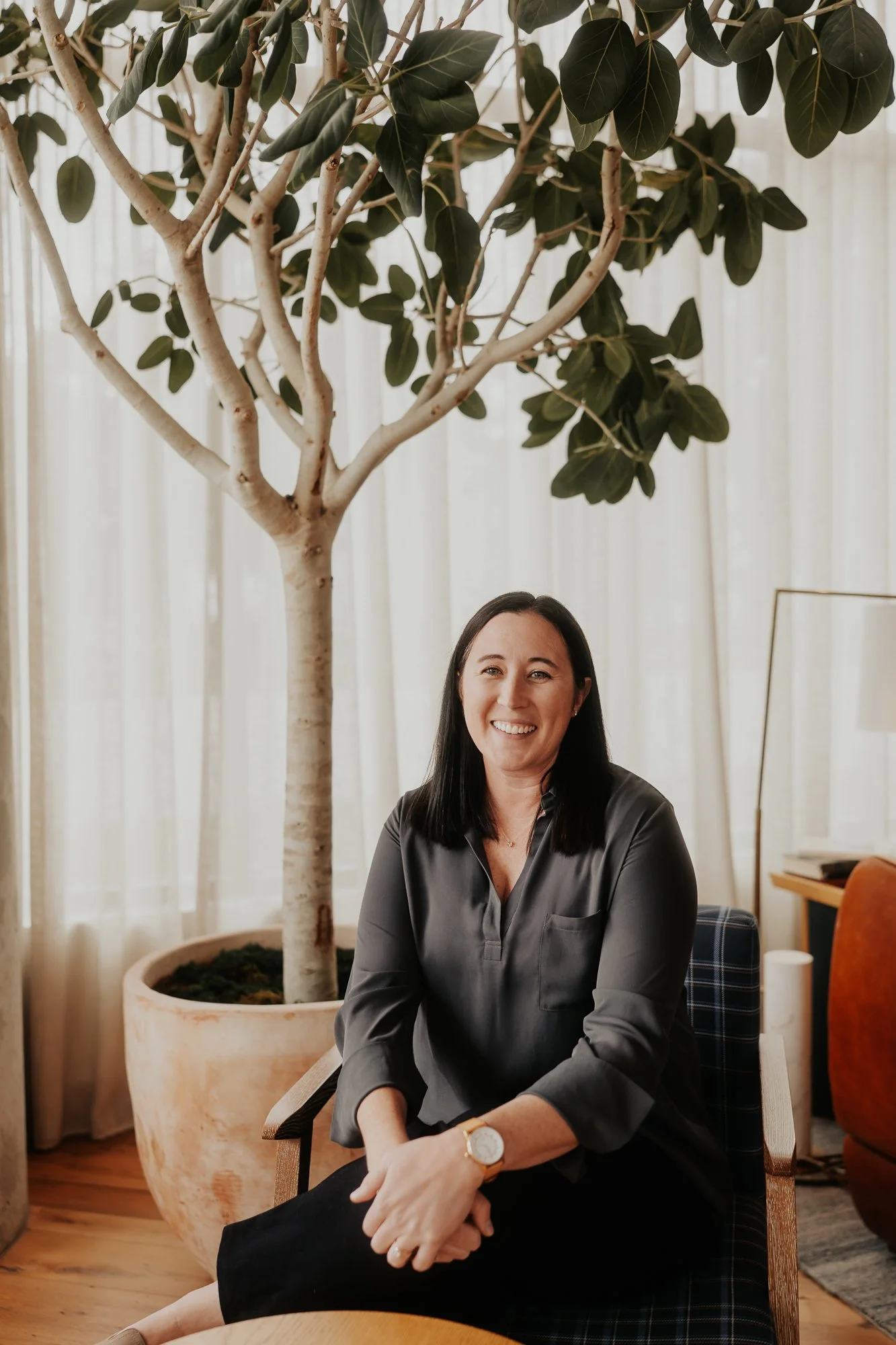 A woman with dark hair smiling, sitting indoors on a chair, in front of a large potted plant with a tree, beige curtains behind, wooden floor, and some furniture in the background.