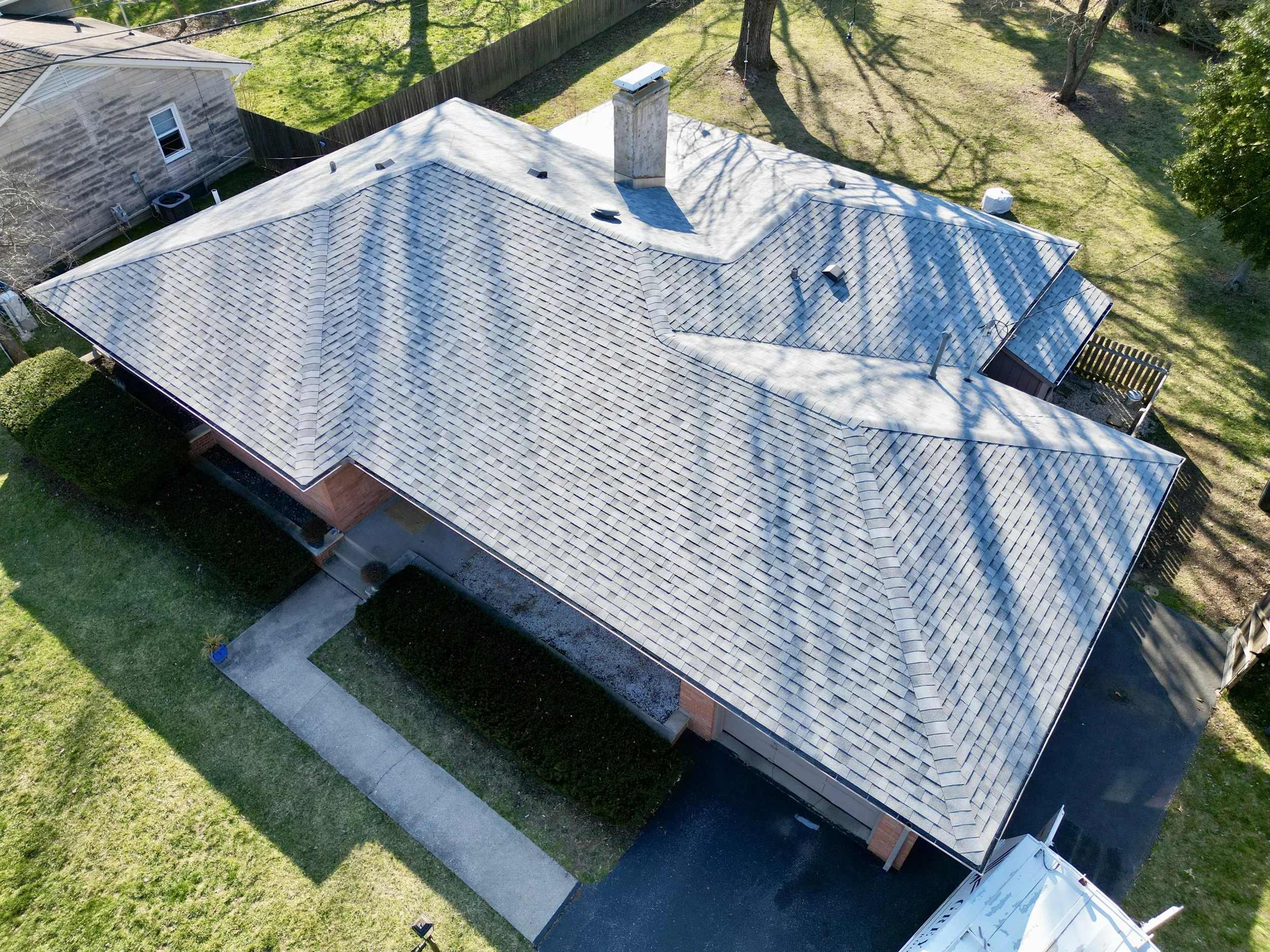 Aerial view of a house with a complex, gray shingled roof, surrounded by a green lawn, trees, and neighboring houses.