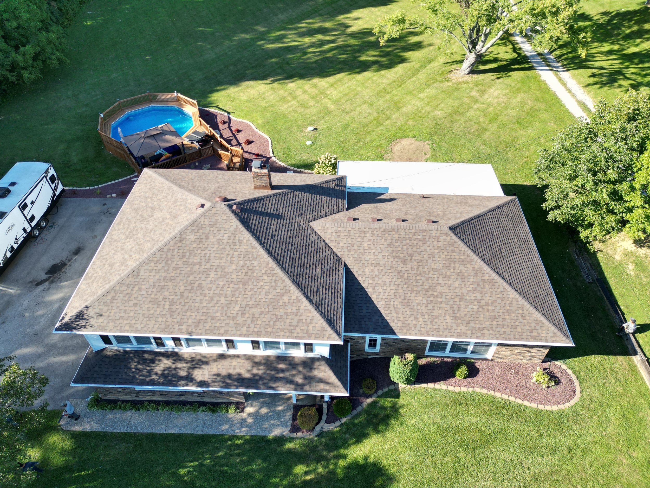 Aerial view of a house with a brown shingle roof, a backyard with a pool and deck, a driveway with a RV, and surrounding green lawns and trees.