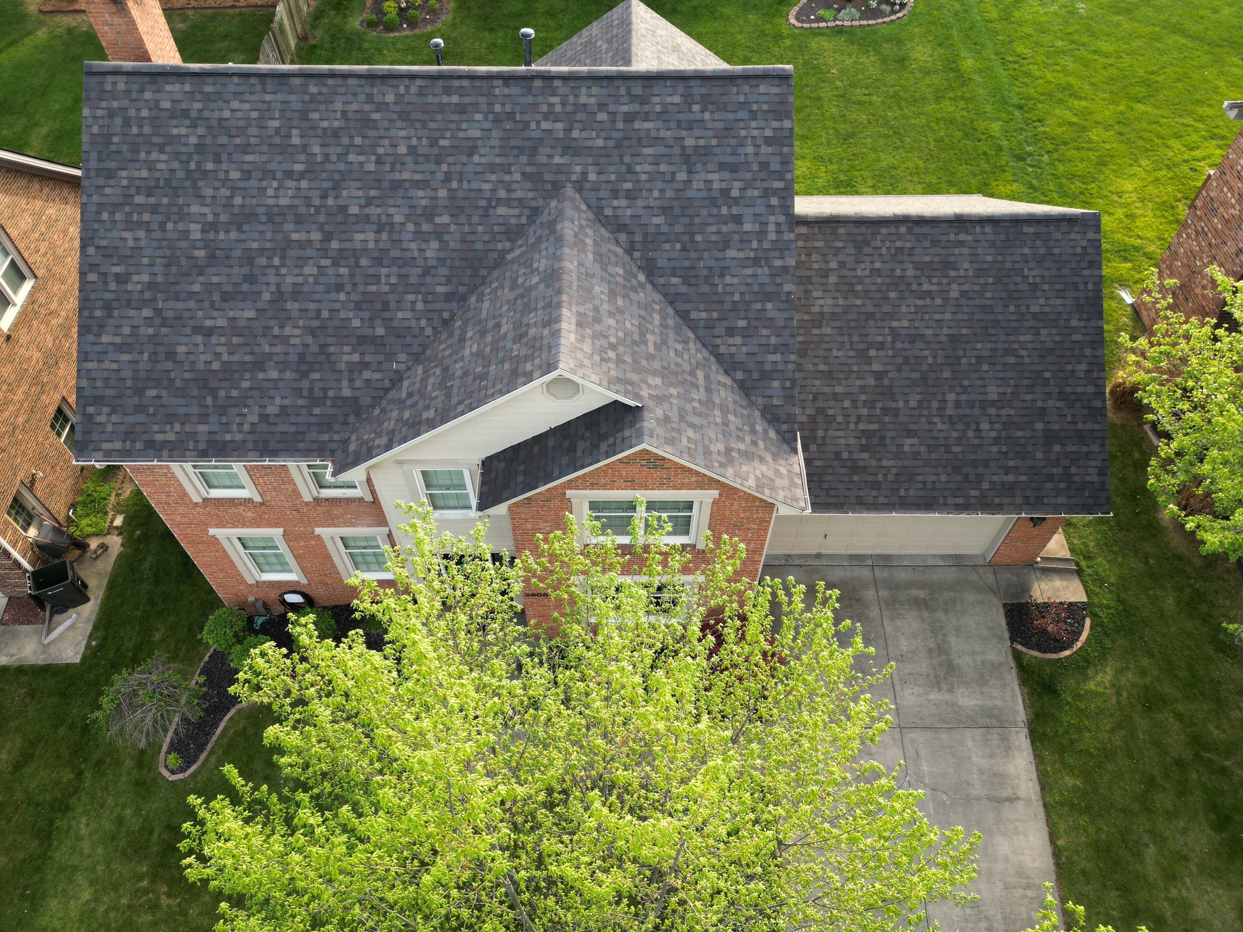 Aerial view of a two-story brick house with a dark shingle roof, surrounded by a green lawn and trees, with a concrete driveway leading to a garage.