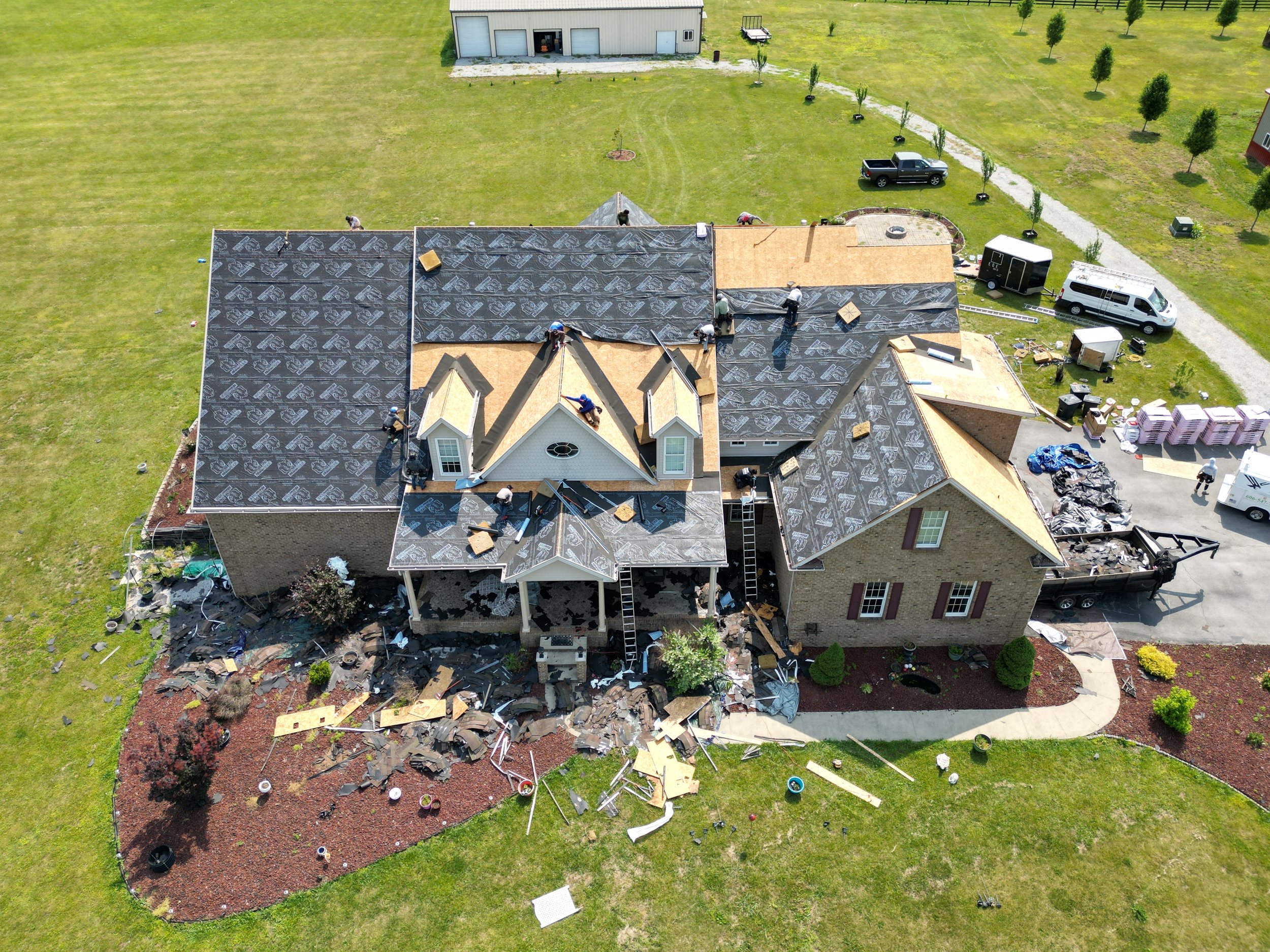 Aerial view of a house under renovation, with workers installing new roofing shingles, surrounded by tools, debris, and construction materials.