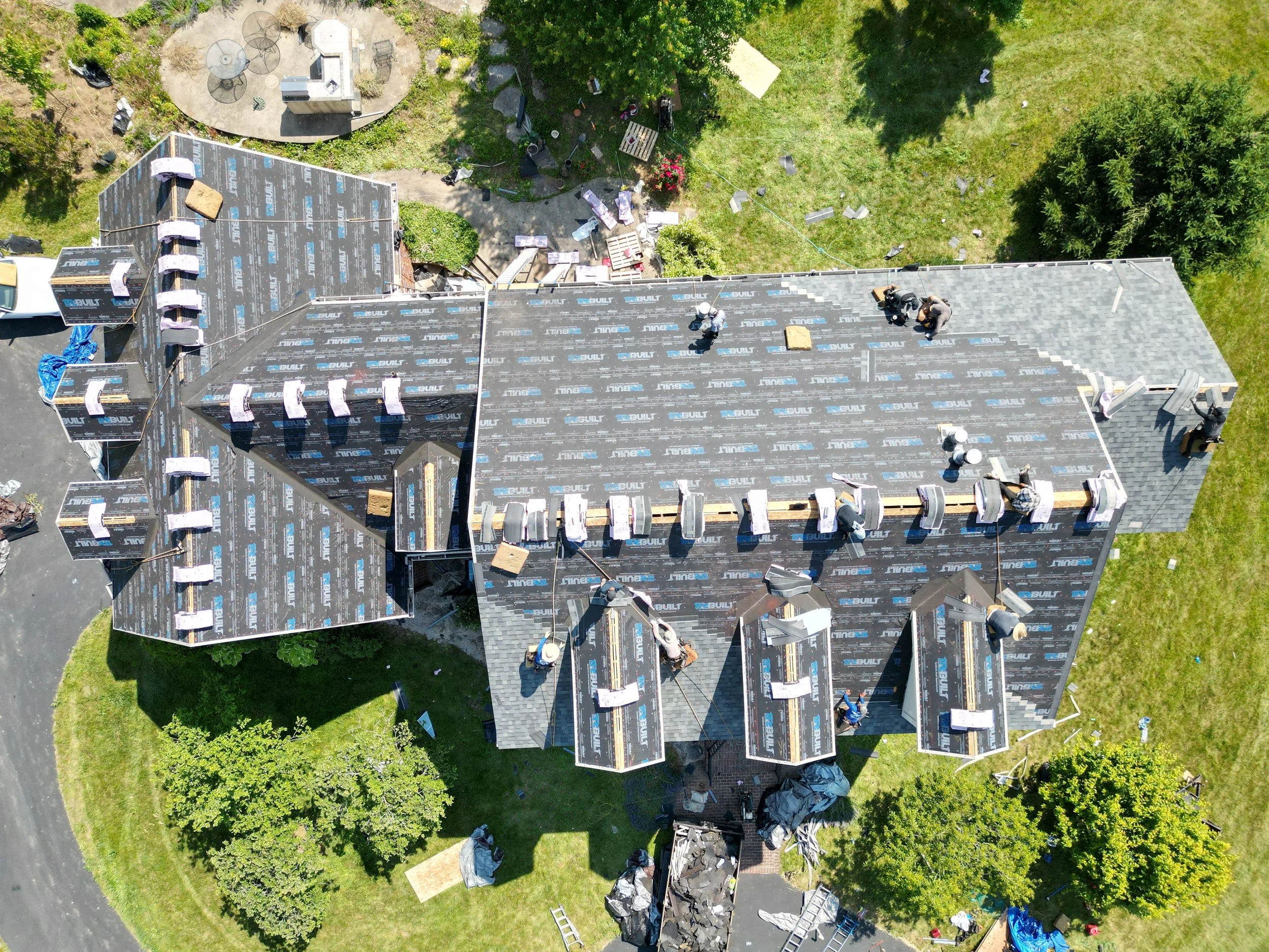 Aerial view of a house under construction with workers installing roofing materials, surrounded by green lawn and trees.