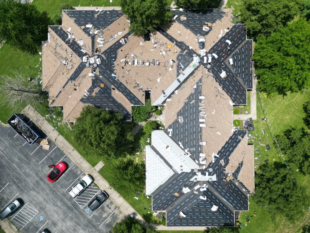 Aerial view of a large residential building with multiple brown roofs, some with missing shingles, surrounded by green trees and a parking lot with several cars.