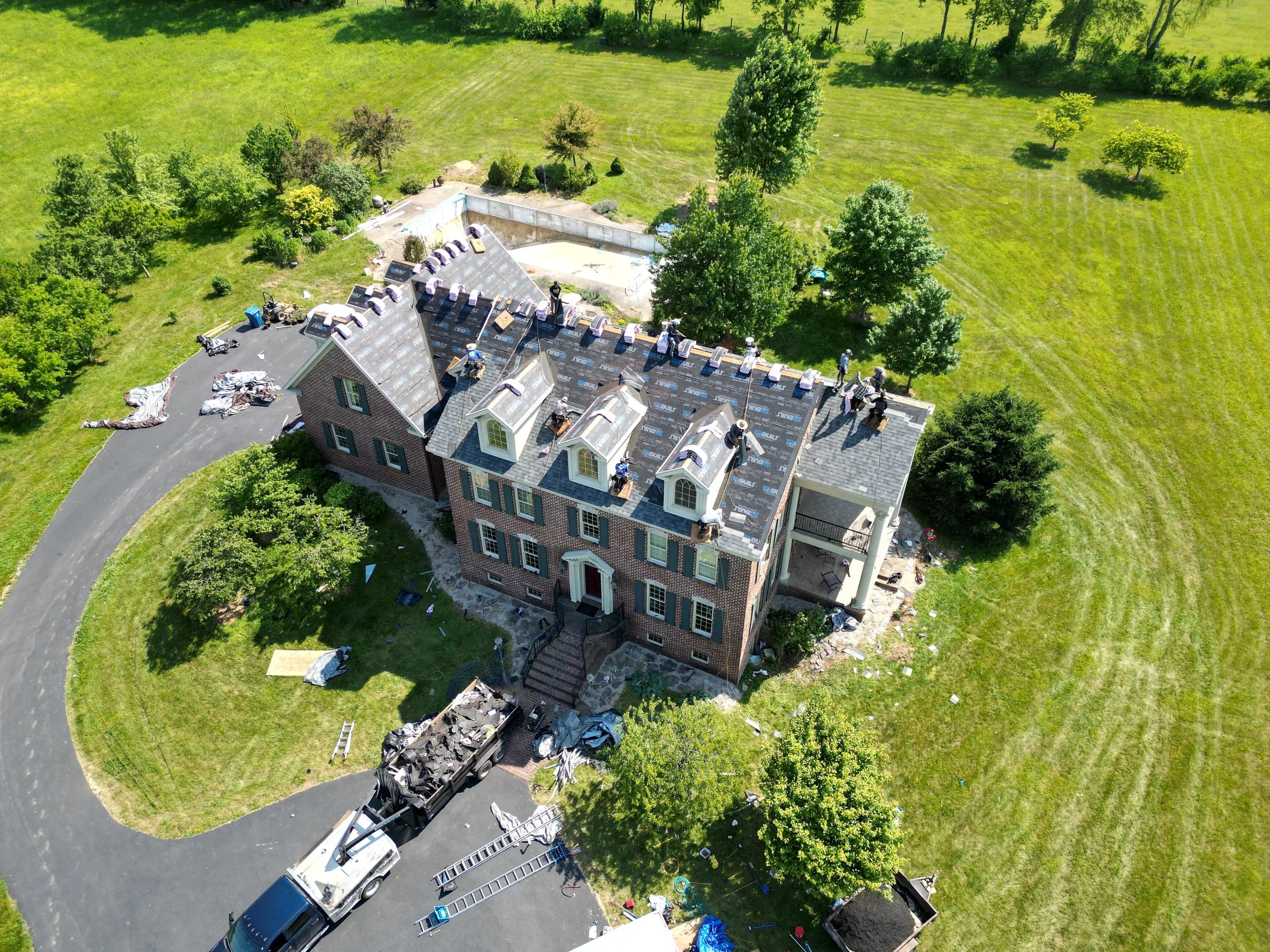 An aerial view of a house under construction with workers on the roof, surrounded by green lawns, trees, and construction equipment.