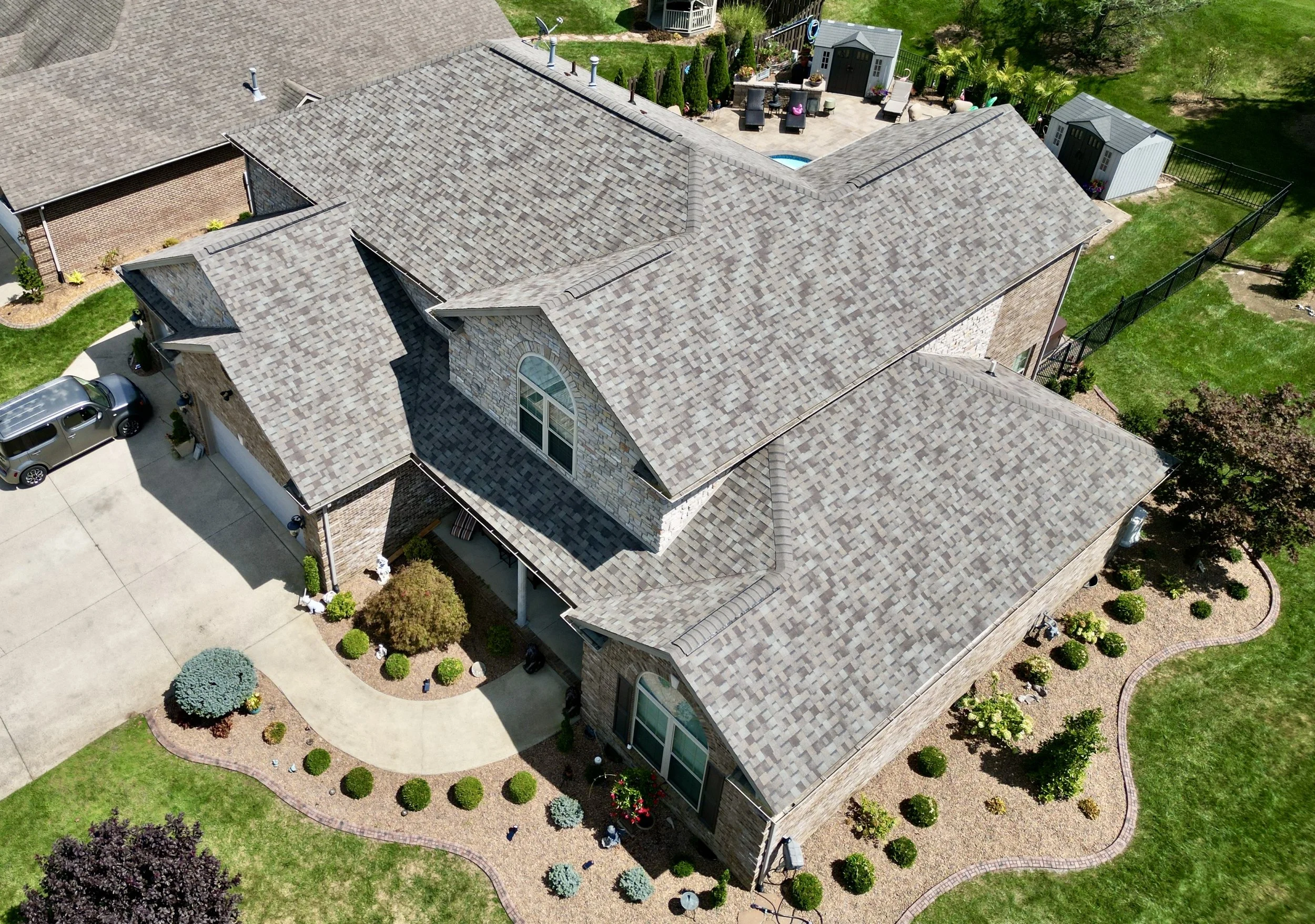Aerial view of a large brick house with a complex gray shingle roof, surrounded by a well-maintained lawn, flower beds, and a concrete driveway with parked cars. Backyard features a patio area with outdoor furniture and a small swimming pool.