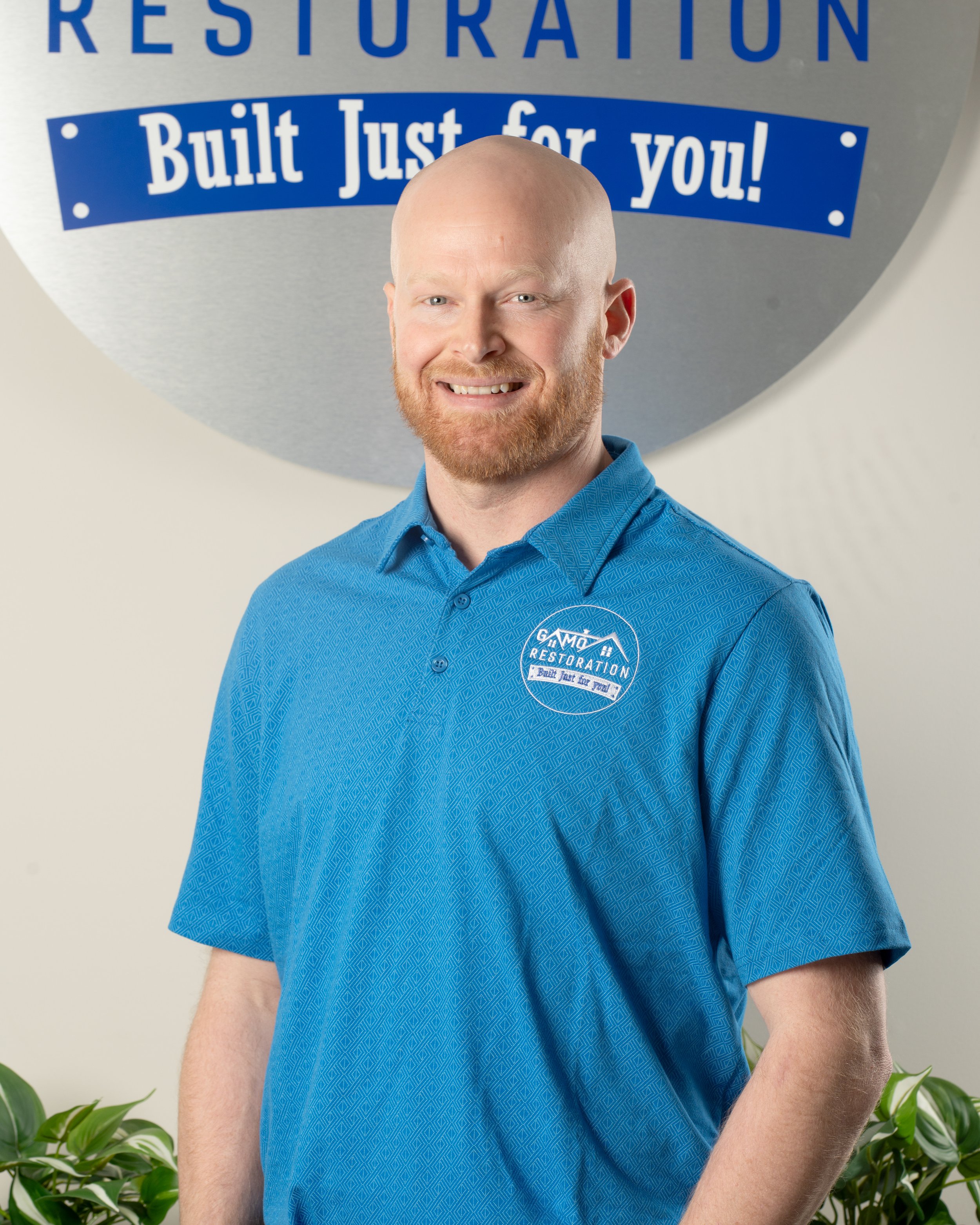 A smiling man with a beard and bald head wearing a blue polo shirt logoed with 'GAM Restorations' stands in front of a large sign that reads 'Restoration Built Just for You!' in blue and white lettering.