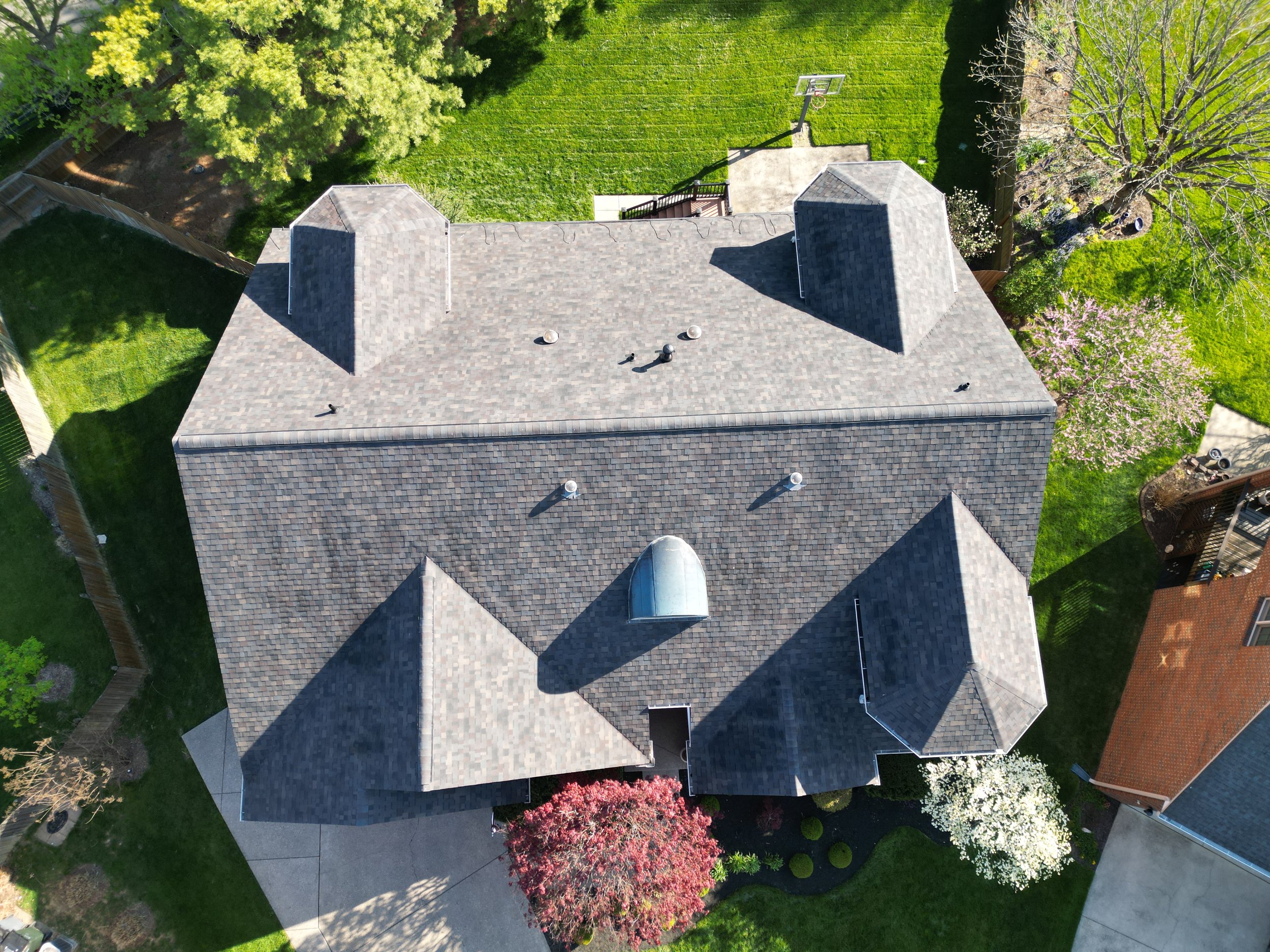 Aerial view of a house with gray roof and multiple gables, surrounded by green lawns and trees, with a basketball hoop and a patio in the backyard.