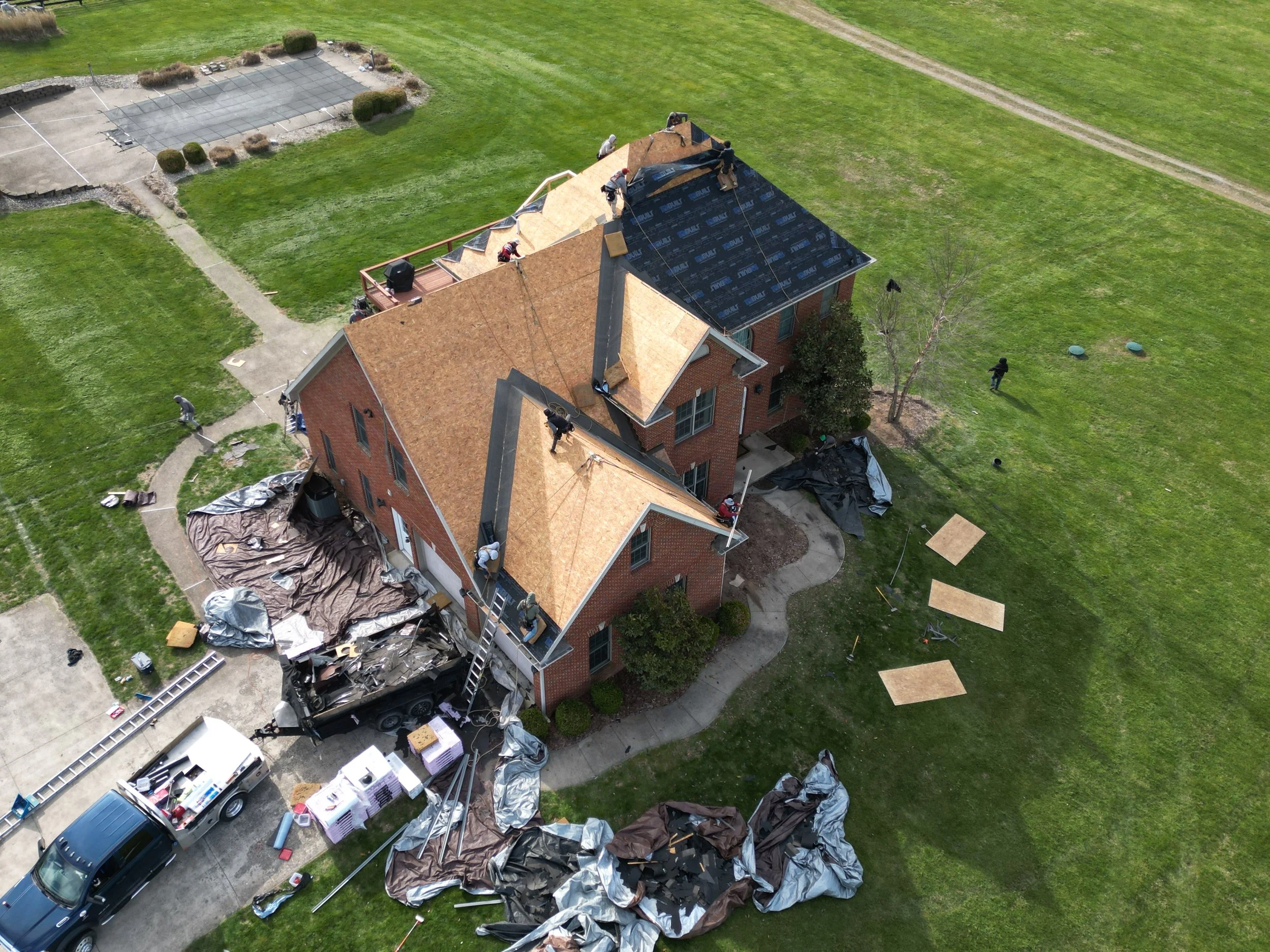 Aerial view of a house under roofing renovation with workers on the roof, roofing materials on the ground, and construction tools and debris around the property.