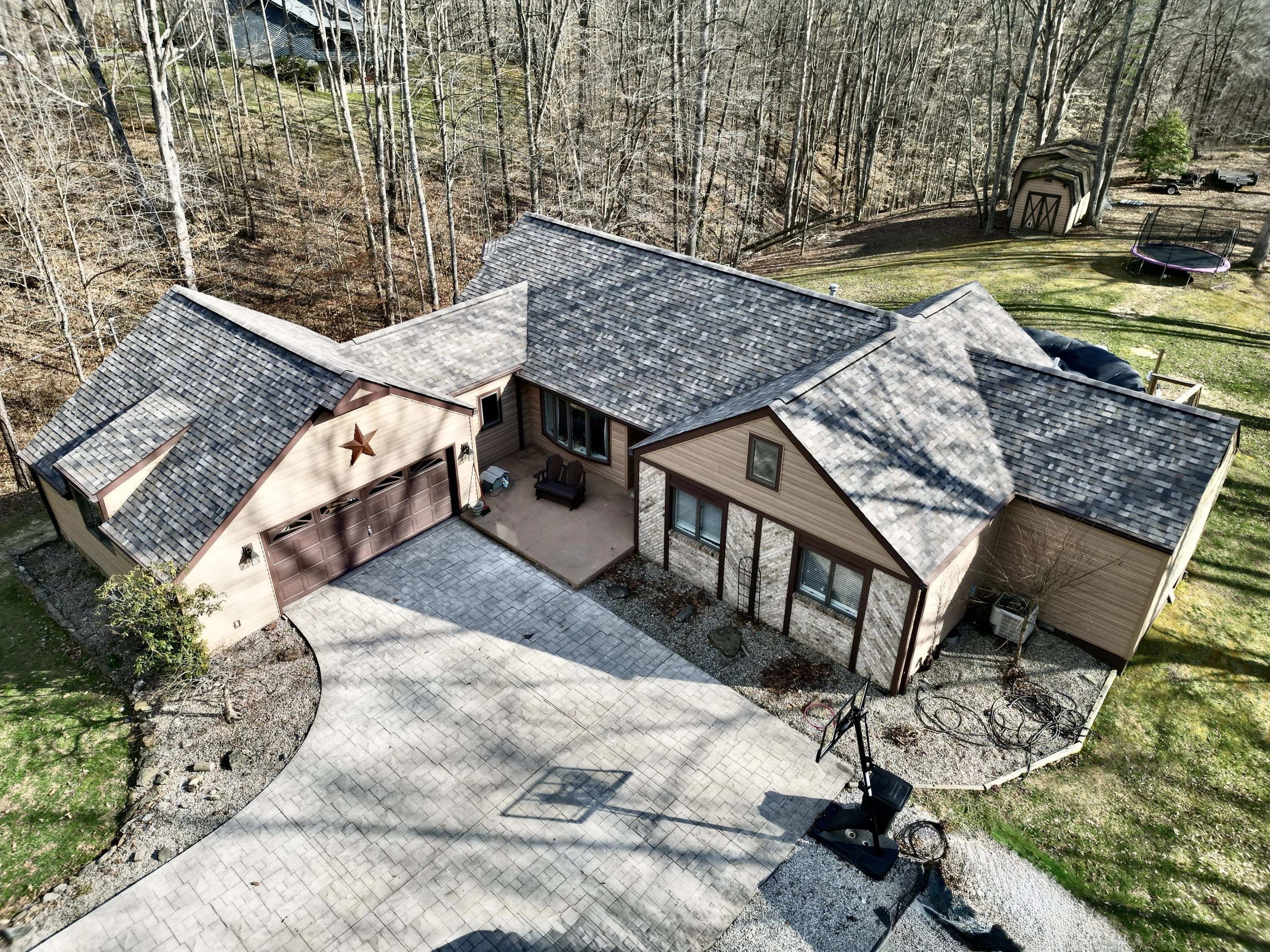 An aerial view of a house with a shingled roof, a driveway, a basketball hoop, a small backyard with a trampoline, a storage shed, and surrounding trees.
