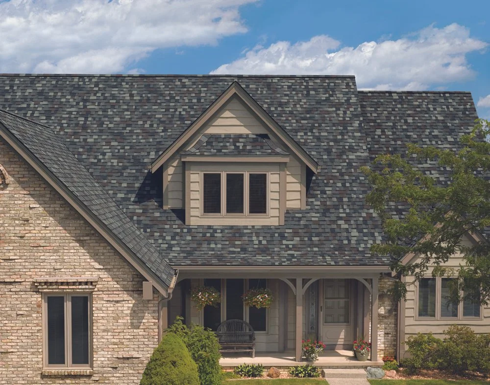 Front view of a house with a brick and siding exterior, gray shingle roof, dormer window, front porch with hanging flower baskets, and surrounding greenery under a partly cloudy sky.
