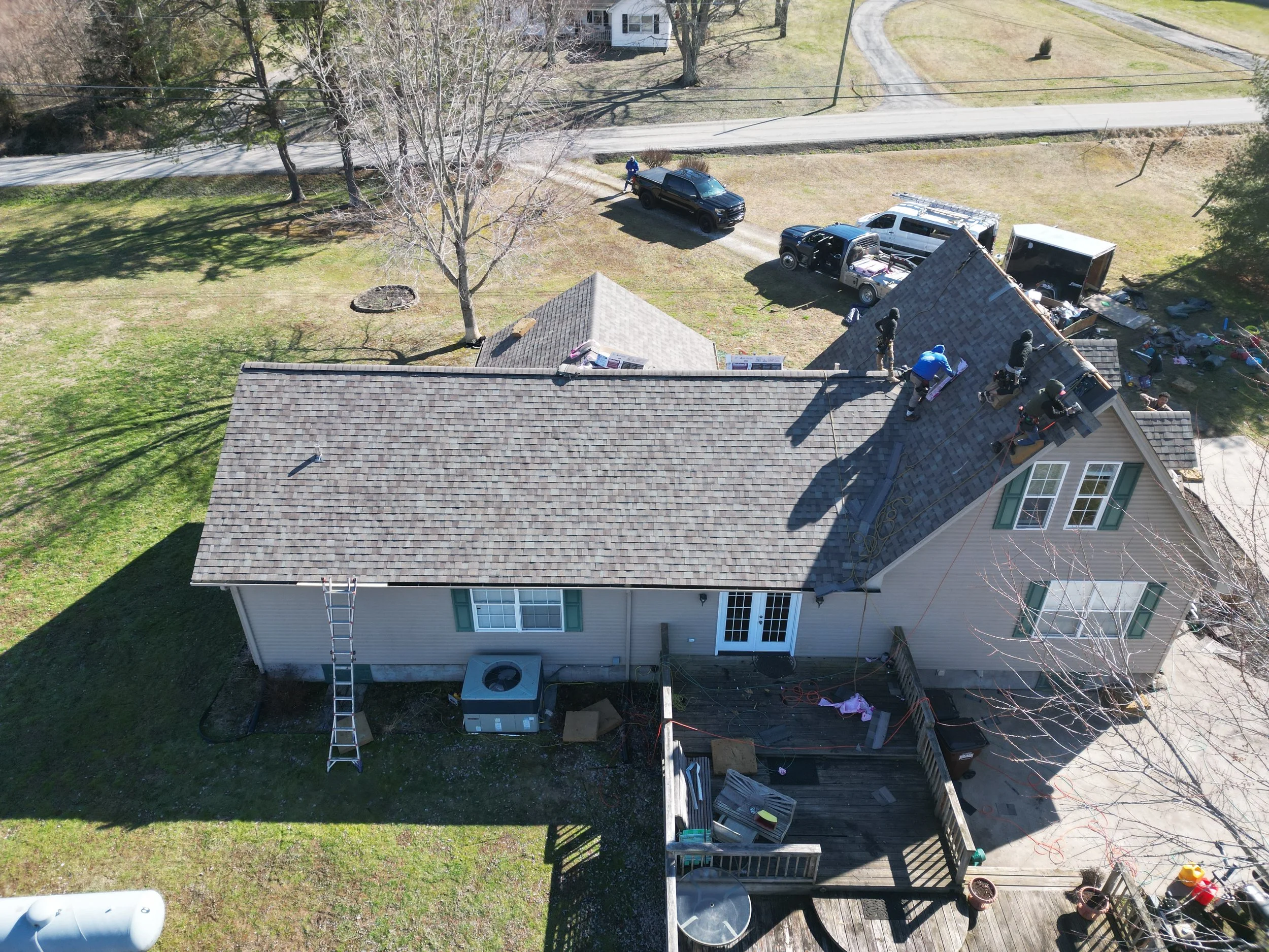 Aerial view of a house with workers replacing the roof shingles on the right side, with several vehicles parked nearby and trees surrounding the house.