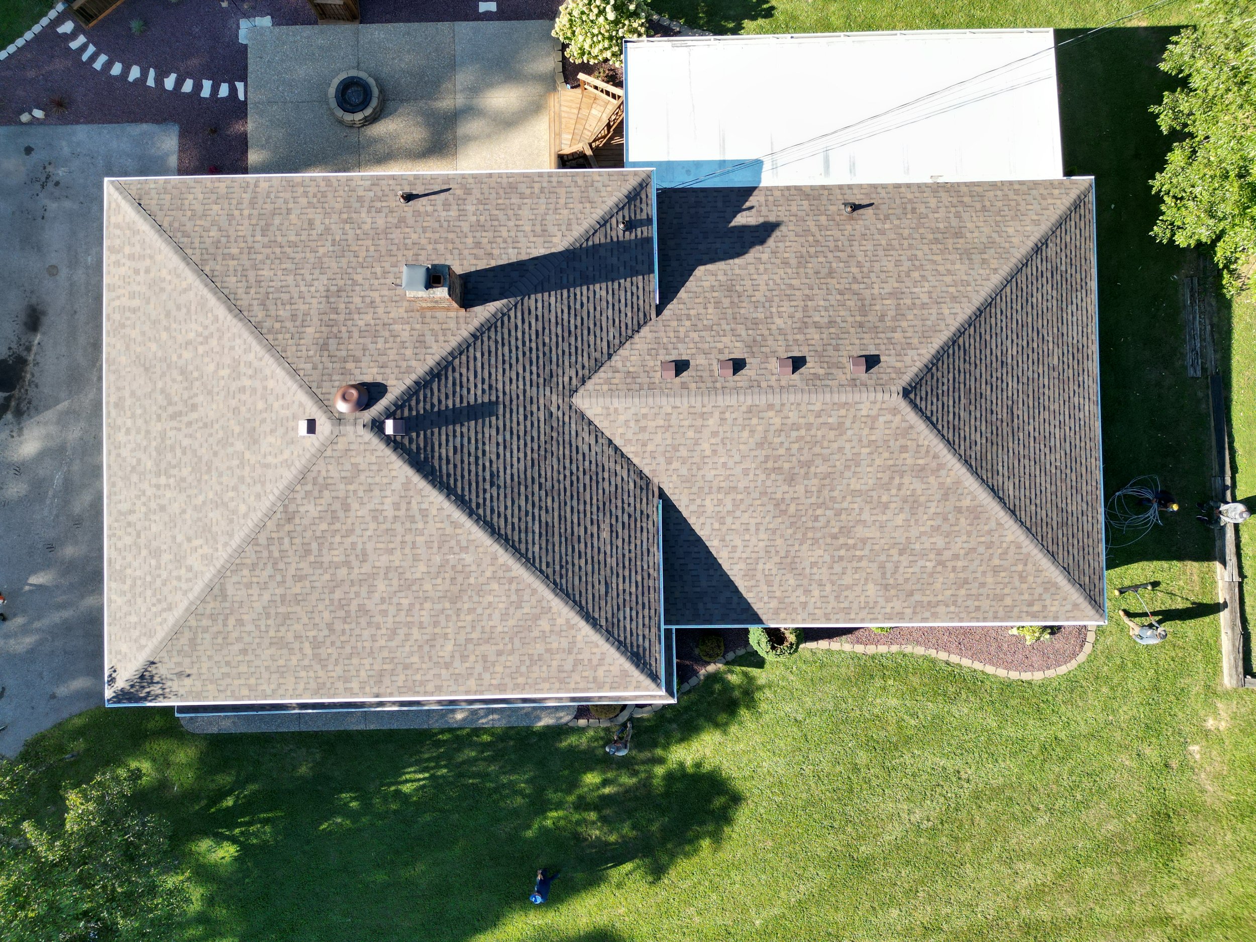 Aerial view of a house with a brown shingle roof, surrounded by a green lawn with some trees, garden beds, and outdoor items, and adjacent to other structures and paved areas.