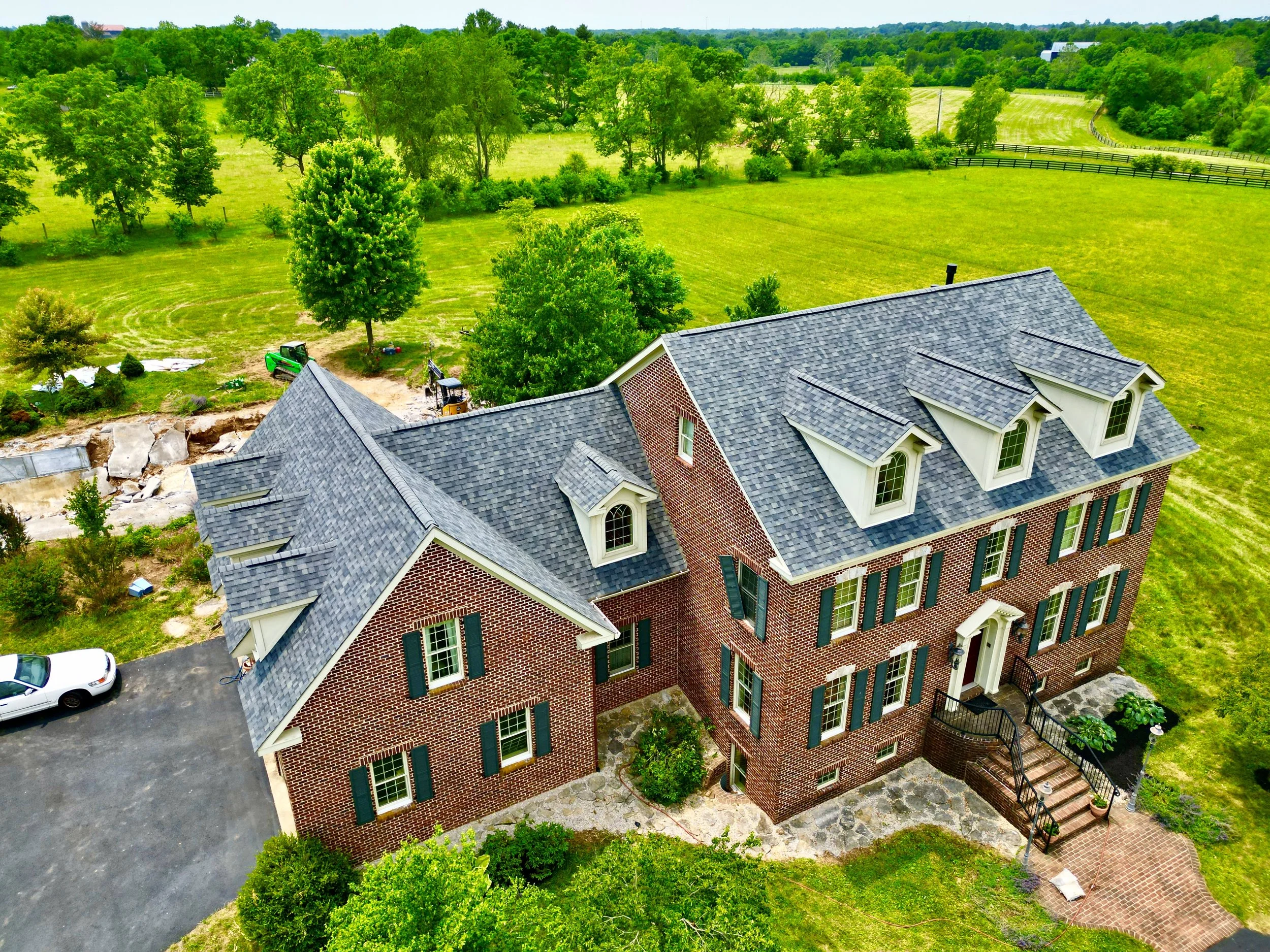 A large brick house with a dark gray shingle roof and green shutters, situated on a grassy, rural landscape with trees and open fields behind it.