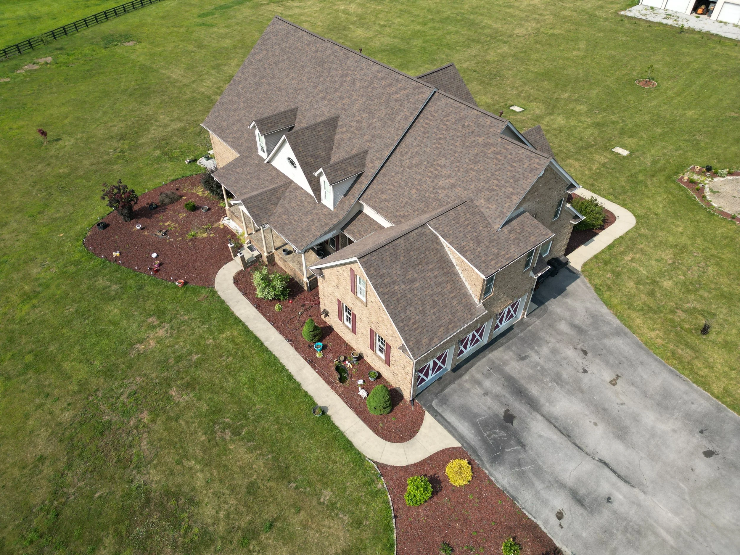 Aerial view of a two-story house with a brown shingled roof, surrounded by a well-maintained lawn, concrete driveway, and landscaped yard with decorative bushes and mulch beds.