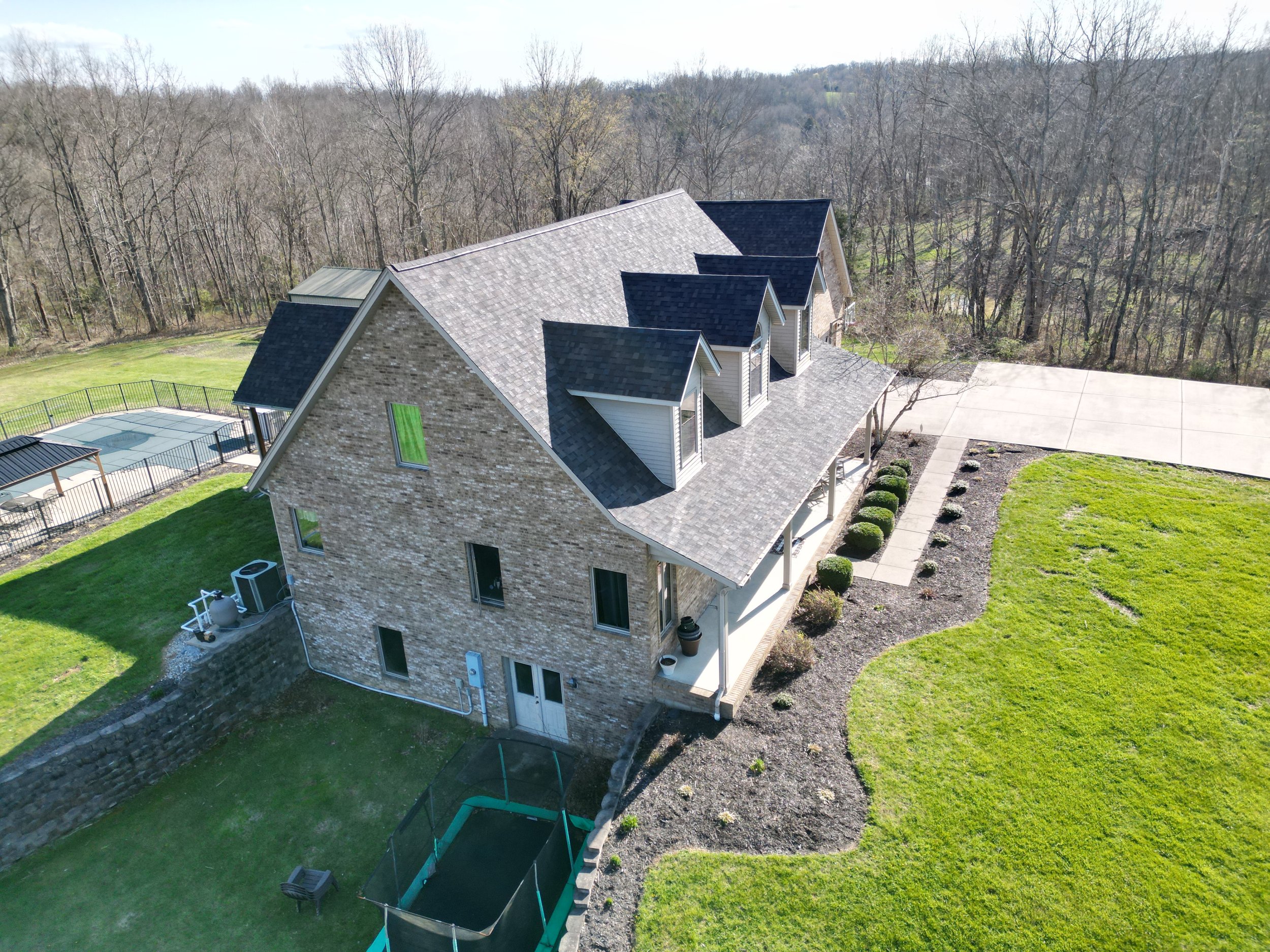 Aerial view of a large house with a brick and siding exterior, multiple gabled roofs, a backyard with a small pool, a patio, a lawn, and wooded area in the distance.