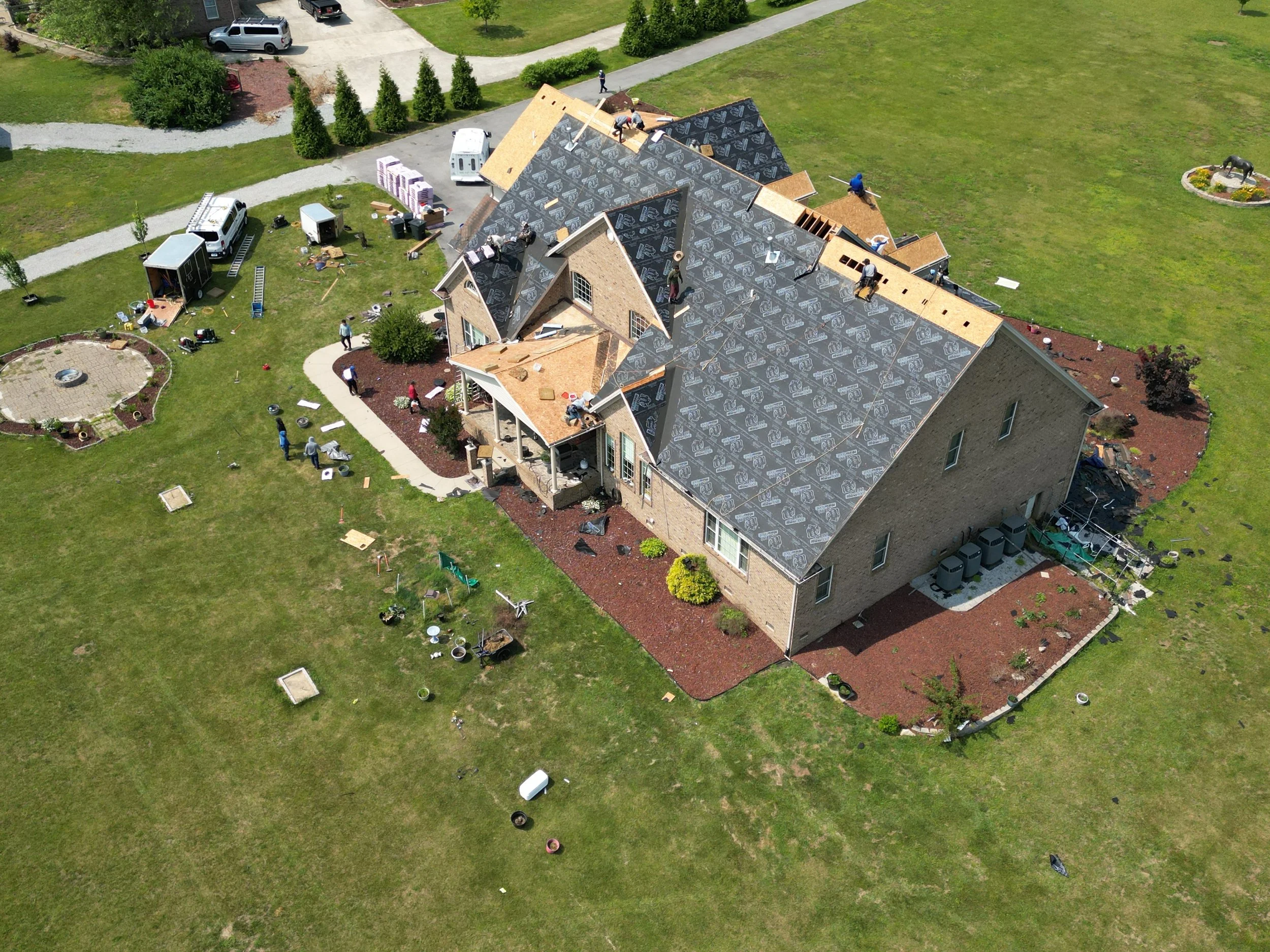 Aerial view of a house under construction with workers installing roofing. The house has a brick exterior, with sections of roof covered in black roofing underlayment and other sections with wooden sheathing. Construction equipment and materials are 