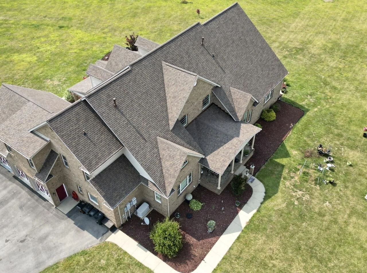 Aerial view of a large residential house with a complex roof design, brick exterior, and front porch surrounded by a landscaped yard with grass and shrubs.