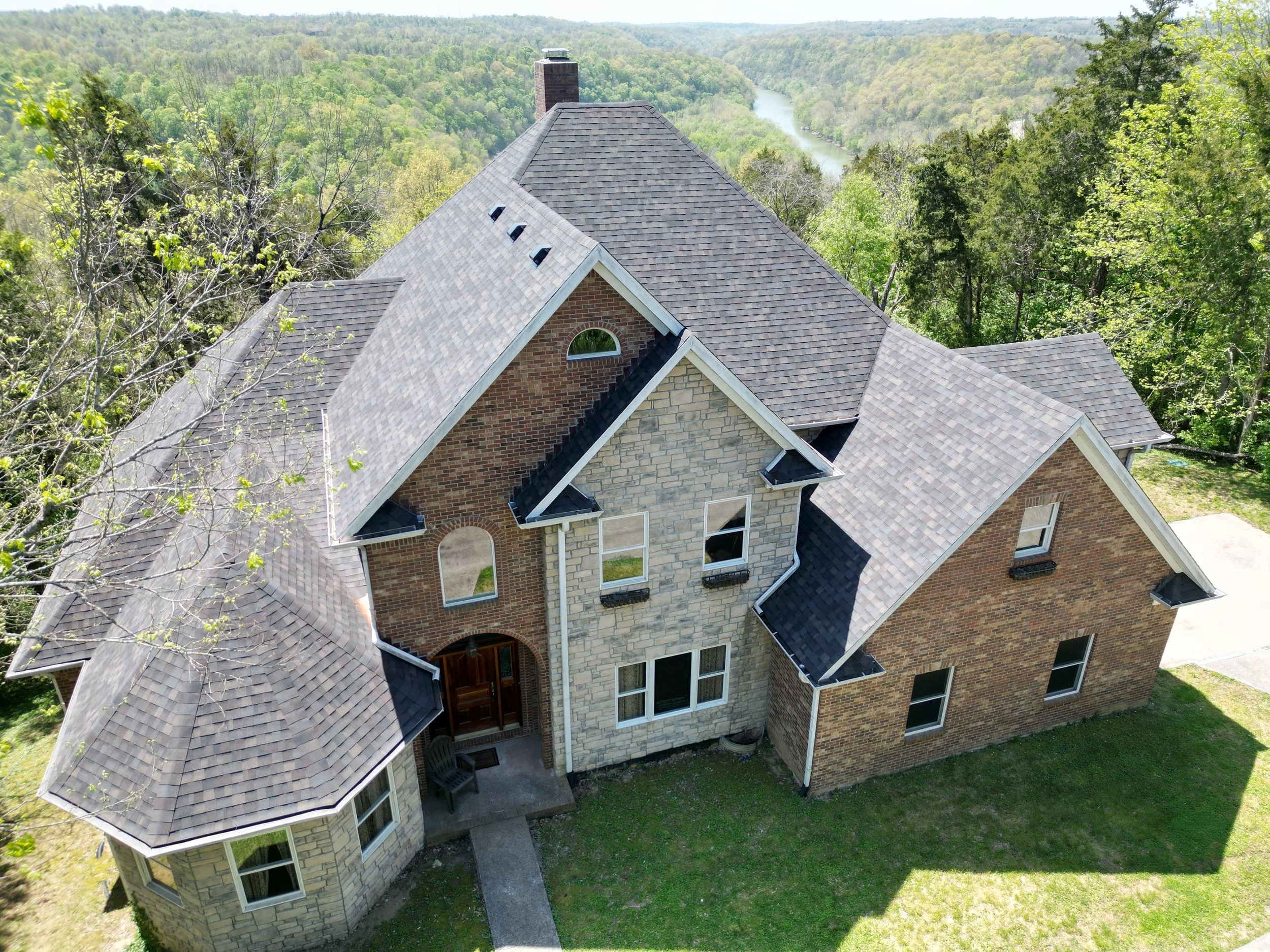 Aerial view of a large two-story house with a brick and stone facade, surrounded by green trees and overlooking a river in the distance.