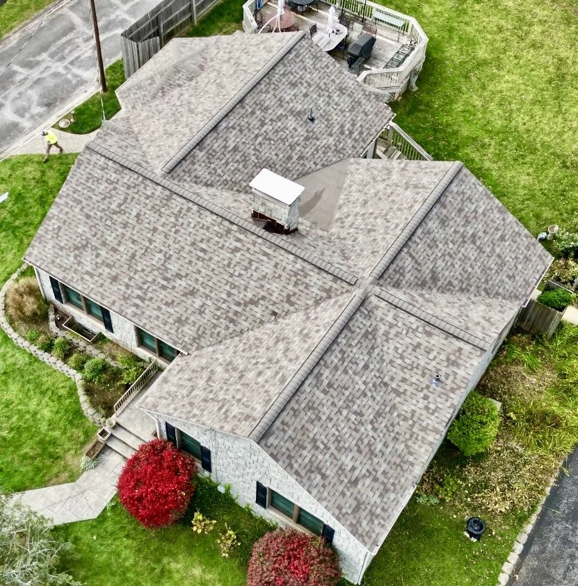 An aerial view of a house with a multi-gabled roof, surrounded by green grass and trees, with part of a deck visible on the back side.
